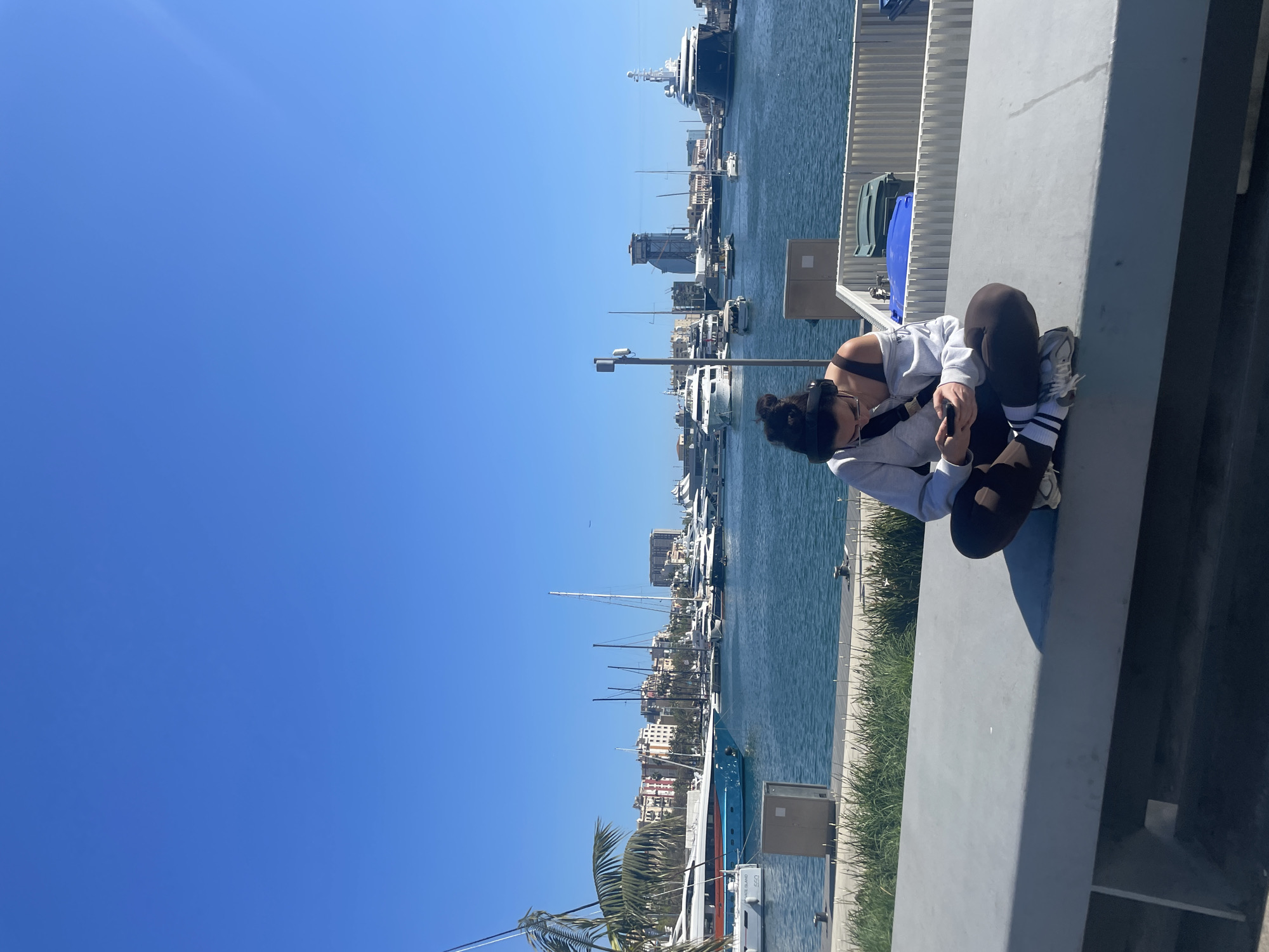 A vertical, medium shot captures a young woman sitting on a low concrete wall in Barcelona, Spain, overlooking the harbor filled with various boats and yachts. The woman is positioned on the right side of the frame. She has dark hair pulled up in a bun, wearing a light gray hoodie, dark leggings, and sneakers with black and white striped socks. Her head is bowed, and she appears to be focused on something in her hands, possibly a mobile phone.
The harbor stretches out behind her, filled with boats ranging from small sailboats to larger yachts and even a large ship further in the background. Buildings line the waterfront beyond the boats. The sky is clear and bright blue. Palm trees are visible at the lower left corner of the frame. The foreground is dominated by the gray concrete wall where the woman sits, with some greenery growing along its base.
