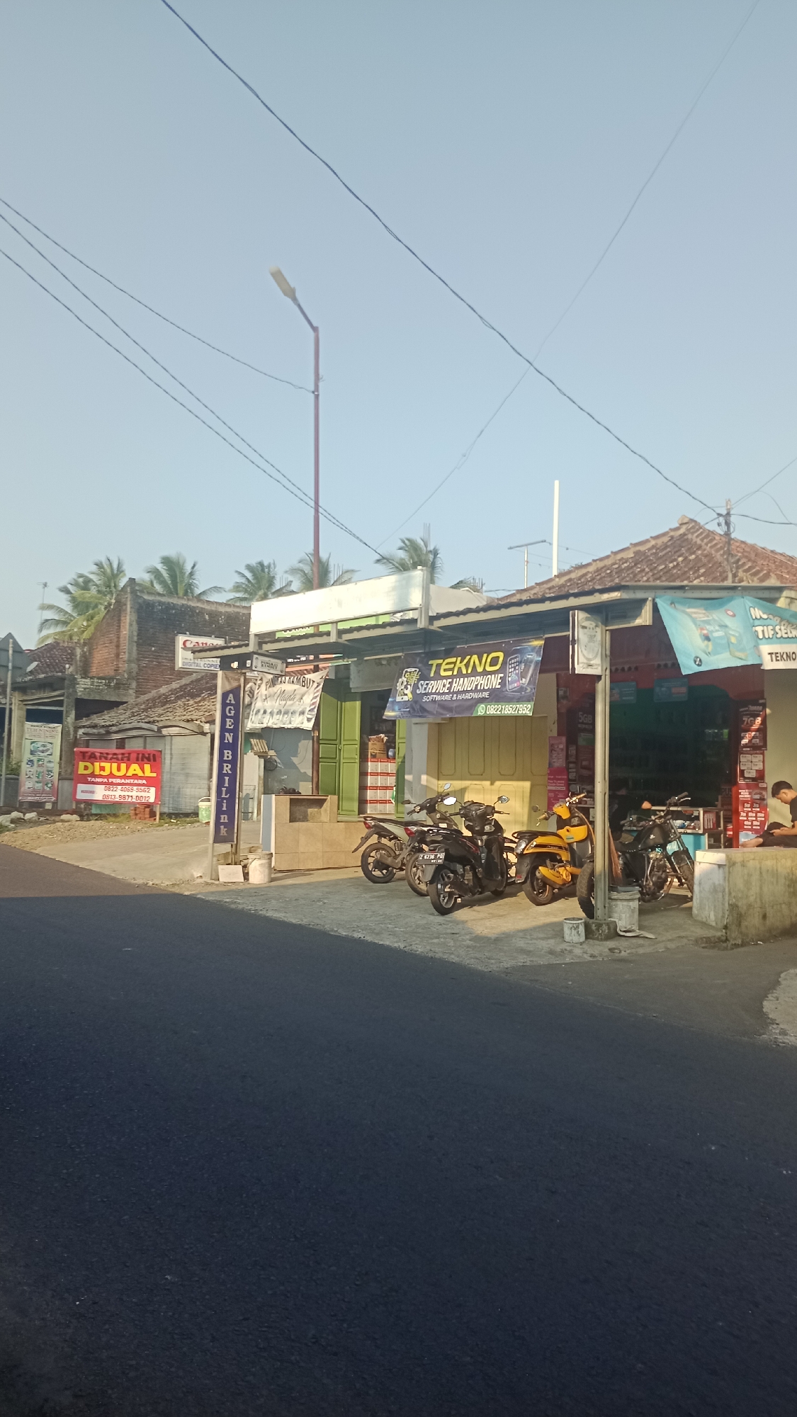 The image shows a street scene in Karyamandala, Indonesia, likely during the daytime with clear skies. On the right side of the street, there is a row of small shops. One shop prominently displays a banner that reads "TEKNO SERVICE HANDPHONE SOFTWARE & HARDWARE" with a phone number. Adjacent to this, another banner mentions "TIF SE...". Several motorcycles are parked in front of these shops, including a yellow scooter and two black motorcycles. A person is seated on a low wall in front of the rightmost shop, appearing to be relaxing or observing.
To the left of the shops, there is a sign for "AGEN BRILINK" and another sign with the words "TANAH INI DIJUAL" (This land is for sale), along with phone numbers. Further to the left, a building has a "Canon" sign, suggesting a business related to photography or printing. Palm trees are visible in the background behind the buildings. The road in the foreground is paved and appears to be asphalt. Power lines are strung across the sky, and a street lamp is visible in the upper left portion of the image. The overall impression is a typical street view of a commercial area in a small Indonesian town.