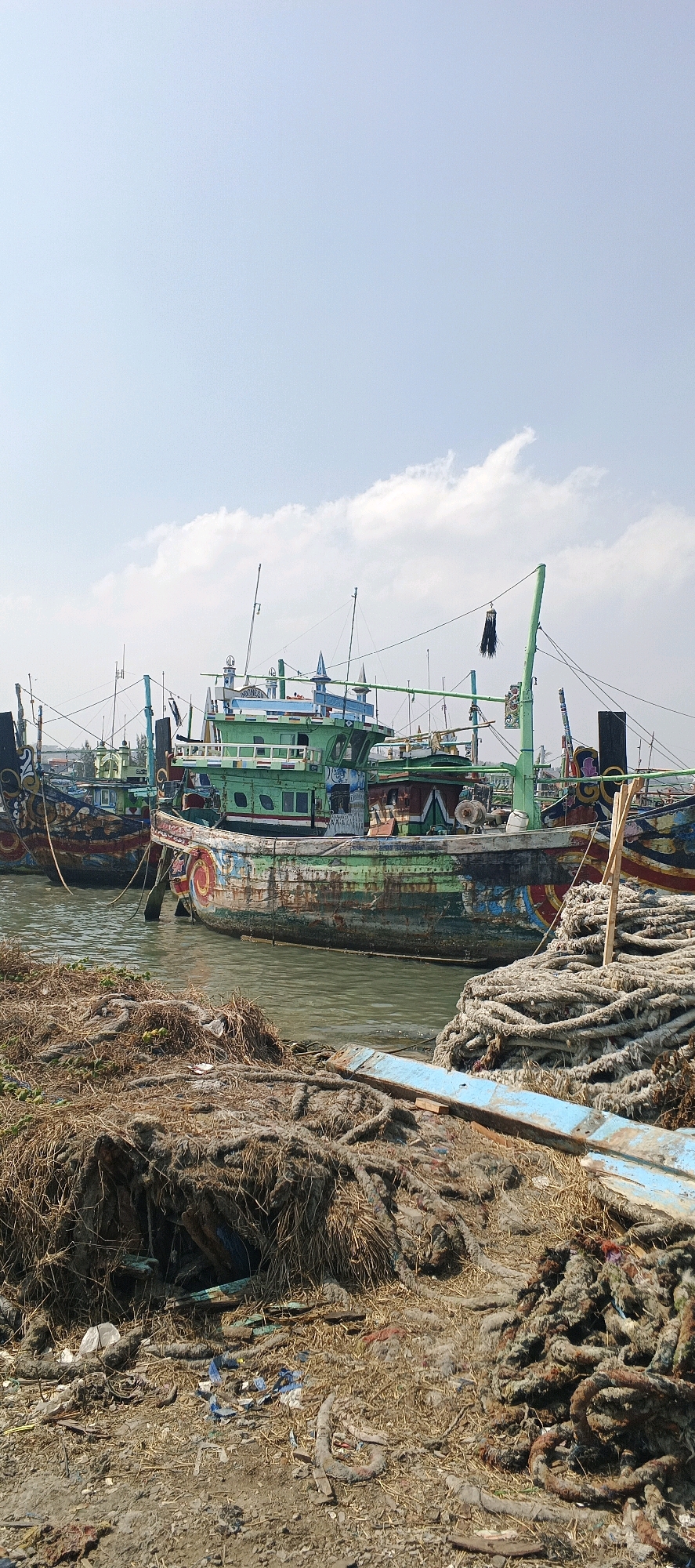 The image depicts a harbor scene in Blimbing, Indonesia, likely a fishing village. In the foreground, a pile of thick, weathered ropes rests on dry, straw-like debris and loose dirt. Some of these ropes appear heavily used and are coiled loosely, while others are tangled with dried vegetation and possibly some plastic waste. Behind the ropes, several fishing boats are moored in the water. The prominent boat in the center is painted predominantly green with colorful decorative patterns on its hull, showing signs of wear and tear. Other boats, also appearing to be fishing vessels, are visible behind it, some with masts and rigging. The water is a murky, greenish-brown. The sky above is clear with a few scattered white clouds. The overall impression is one of a working harbor, possibly during a period of rest or preparation for fishing.