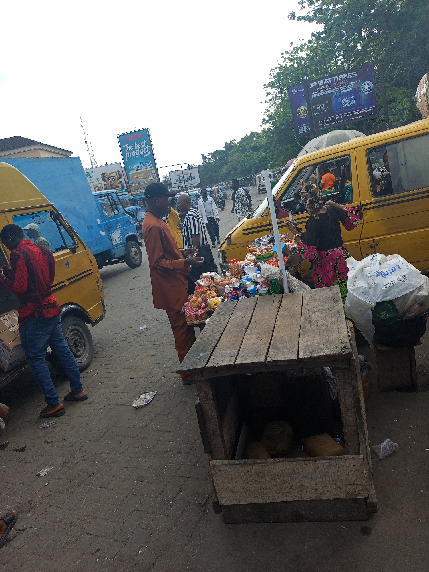 The image depicts a busy street market scene in Satellite Town, Nigeria, during daytime under an overcast sky. The setting is an urban environment with vehicular and pedestrian traffic.
In the foreground, a wooden market stall, constructed from crates, holds various packaged snacks, bottled drinks, eggs, and bread. A woman, likely a vendor, wearing a colorful patterned dress and headscarf, stands behind the stall, appearing to arrange or attend to the goods. To her left, a man in an orange traditional outfit observes the stall, with another man in a striped shirt and black cap standing nearby.
Several vehicles are present. A prominent yellow mini-van, commonly used for public transport, is parked near the vendor's stall, with people visible inside. Further to the left, another yellow van is being loaded or unloaded by a man in a red patterned shirt and blue jeans. A blue truck and other vehicles are seen further down the street, indicating a bustling thoroughfare. People are also walking in the background.
Notable details include the paved ground, scattered litter, and large billboards rising above the street. One billboard advertises "The best product." Another prominent billboard for "JP BATTERIES" states "NOW IN ASPANDA," offering "MORE POWER, LONGER LIFE, GUARANTEED PERFORMANCE," with "12 MONTHS WARRANTY" and "15 MONTHS WARRANTY" seals, a phone number "0814 877 7630," and website "WWW.JPCHRIS.COM," along with an address mentioning "ASPANDA TRADE FAIR." A white plastic bag near the stall has the partially visible text "Lotrec." The overall atmosphere is one of active commerce and daily urban life.