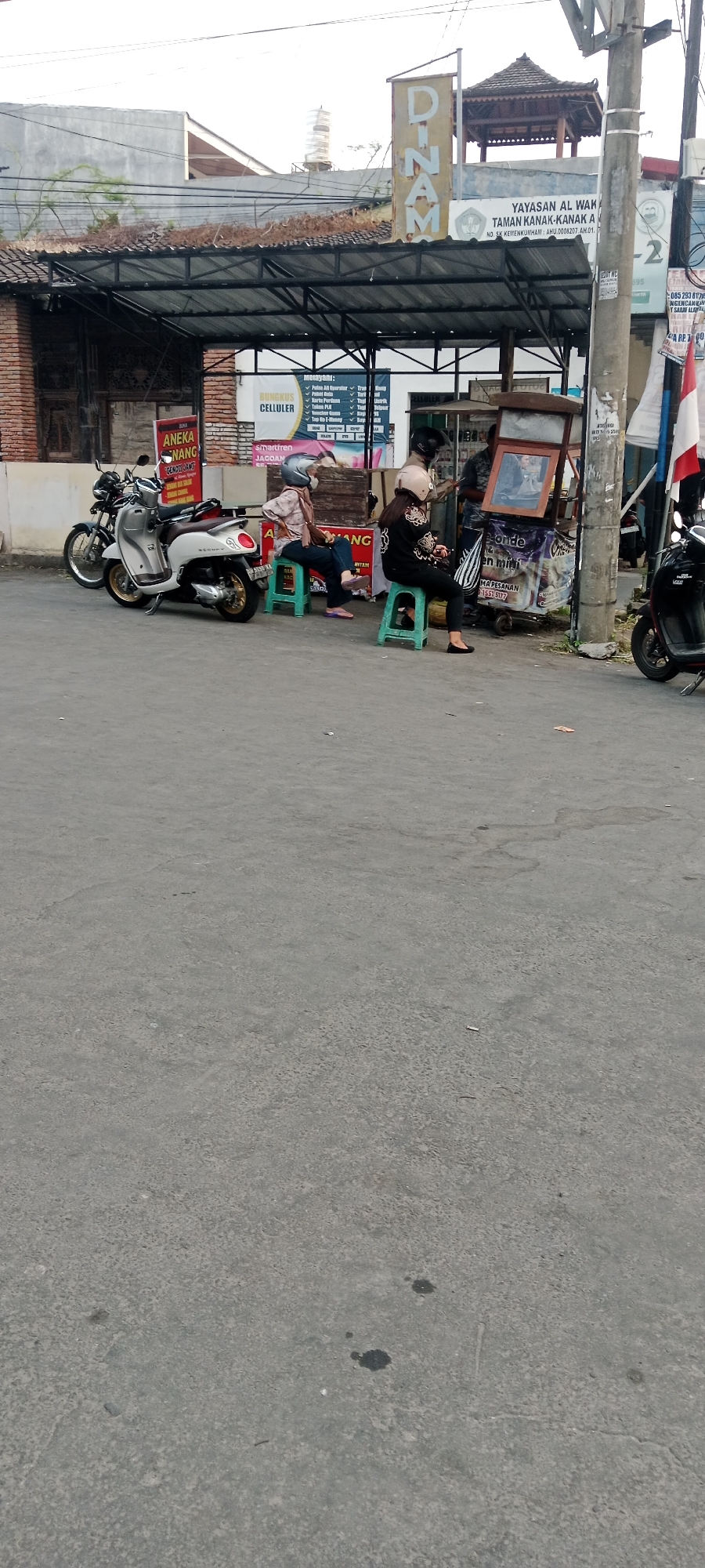 In Maguwoharjo, Indonesia, the image captures a street scene with two women sitting on green plastic stools near a small food cart. The woman on the left wears a light-colored patterned shirt and dark pants, with a silver helmet. The woman on the right is dressed in a black patterned long-sleeved shirt and black pants, also wearing a helmet. They appear to be waiting or resting.
In front of them, a white scooter is parked, along with a dark-colored motorcycle. Behind the women, a stall displays various items, including what appears to be a framed picture or advertisement. A sign above the stall reads "BUNGKUS CELLULER". Further in the background, a building with a brown tiled roof is visible, and a large sign with the word "DINAM" is mounted high on a pole. The ground is paved with asphalt, showing signs of wear and some scattered debris. The overall atmosphere suggests a typical street-side business operation in a local Indonesian town.