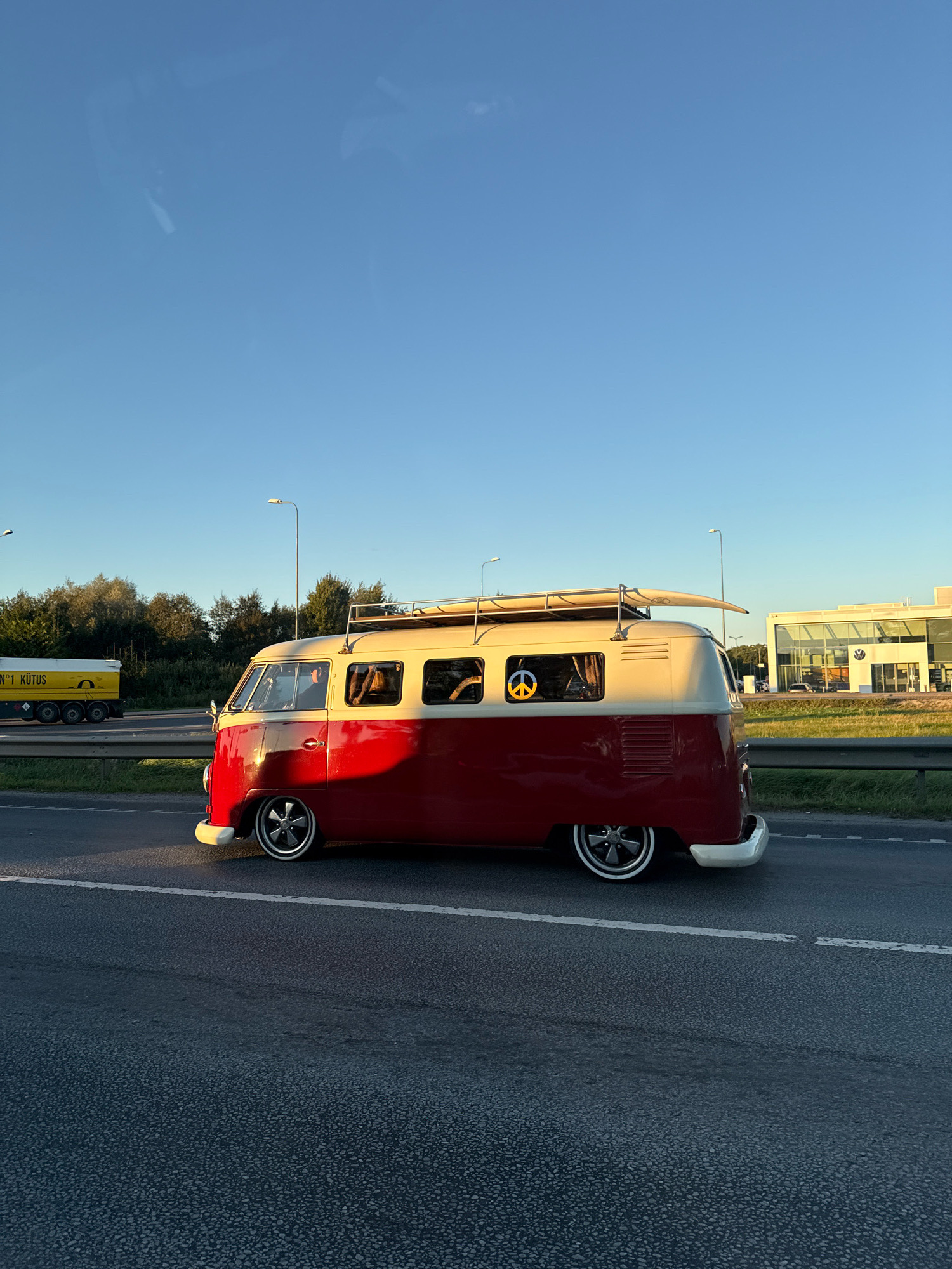 A red and cream colored Volkswagen Type 2 van is captured in a side profile on a highway in Peetri alevik, Estonia. The van, which appears to be a classic model, has a surfboard secured to its roof rack. A prominent peace sign sticker is visible in the passenger window. In the background, a yellow tanker truck with text "N°1 KÜTUS" is on the left side of the highway, and a modern building with a Volkswagen logo is on the right. The sky is clear and blue, indicating daylight, and the sun casts a warm glow on the van. The road is asphalt, with clear lane markings. The environment suggests an urban or suburban setting, likely during the daytime.