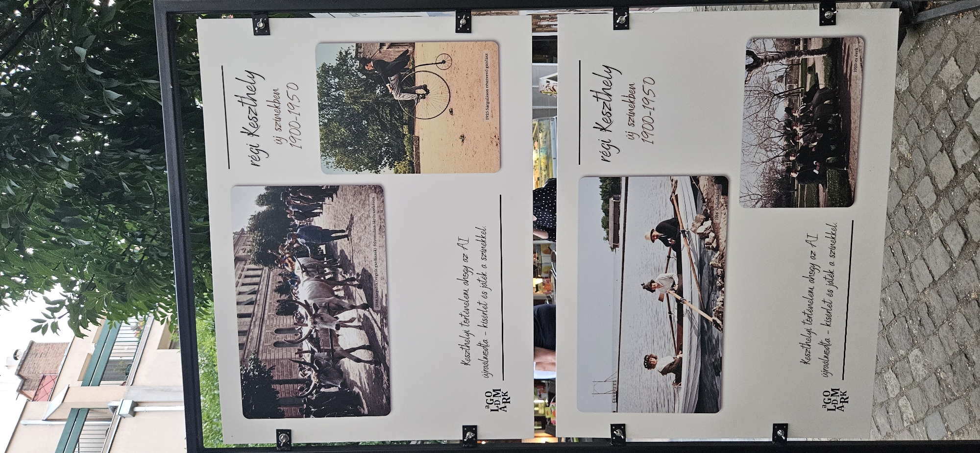 The image displays a panel from an outdoor exhibition in Keszthely, Hungary, featuring historical photographs of the city from the period 1900-1950. The panel is divided into two sections, each with a title "régi Keszthely új színekben 1900-1950" (Old Keszthely in new colors 1900-1950).
The upper section contains two photographs. The photograph on the left, captioned "1938 Sárgulók (végzős gazdászok) felvonulása ökrös szekéren" (1938 Parade of Sárgulók (graduating farmers) on ox-drawn carts), shows a procession of people with oxen pulling carts down a street lined with buildings. The photograph on the right, captioned "1925 Sárguláson résztvevő gazdász" (1925 Farmer participating in Sárgulás), depicts a man riding a penny-farthing bicycle.
The lower section also contains two photographs. The photograph on the left, captioned "1914 Csónakázók" (1914 Boaters), shows three people in a rowboat on a body of water. The photograph on the right, captioned "1910-es évek" (1910s), shows a group of people, likely men, sitting on a bench in an outdoor setting, possibly a park.
Below each section of photographs, there is Hungarian text. The text in the upper section reads: "Keszthelyi történelem ahogy az AI újraalmodta - kísérlet és játék a színekkel." (Keszthely's history as dreamed up by AI - experiment and play with colors.) A logo with "AGOLDMARK" is present on the left side of this text.
The exhibition panel is mounted on a black metal stand and is set up outdoors, with trees and a building visible in the background. The ground appears to be paved with cobblestones.
