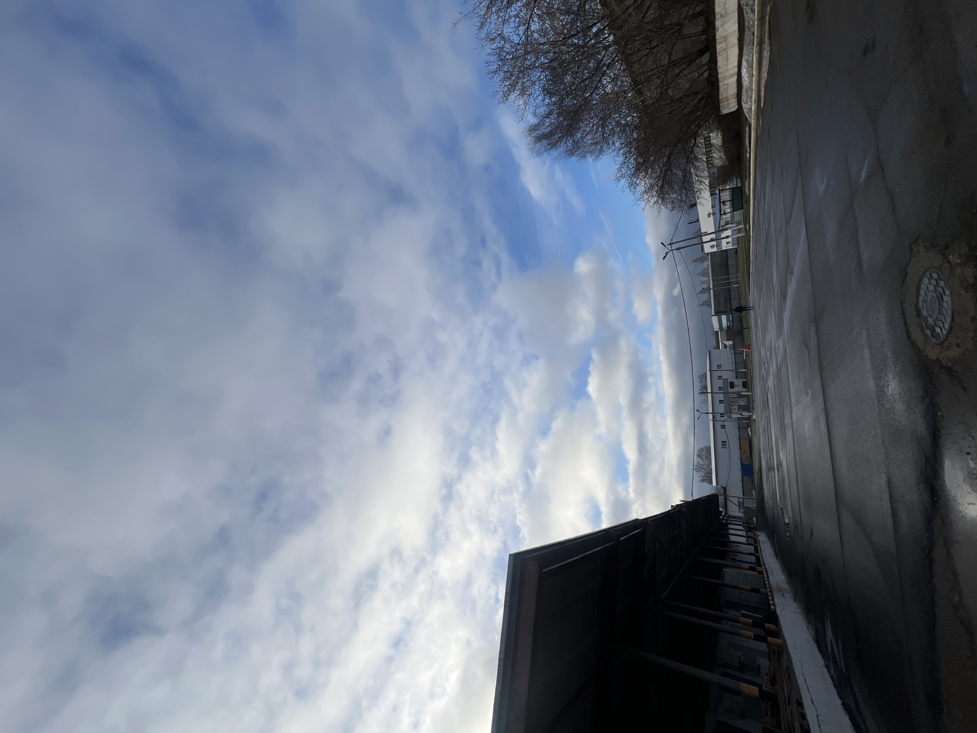 The image captures an outdoor scene, likely a street or an industrial/commercial area, under a dynamic sky. The perspective looks towards a horizon line with buildings and trees.
The sky occupies a significant portion of the frame, filled with a mix of white and light grey clouds against patches of blue sky. Some clouds are soft and wispy, while others are more defined and billowy, suggesting varying atmospheric conditions. Sunlight appears to be breaking through, illuminating some of the cloud formations.
In the lower left foreground, a dark, angular building structure or roof is partially visible, casting a strong silhouette against the brighter sky. To the right, a wide, wet asphalt or paved surface stretches into the midground, reflecting light from the sky and indicating recent rain or snowmelt. A circular metal manhole cover with a patterned surface is distinctly visible on this wet pavement in the lower right.
Along the horizon line, several low-rise buildings, predominantly white or light-colored, are scattered. Bare, deciduous trees with intricate branches stand above these structures, suggesting late winter or early spring. Utility poles with overhead wires are also present in the mid-distance, further defining the urban or semi-industrial setting. A small, dark, indistinct figure can be faintly observed in the distance near the buildings and utility poles, but no specific activity is discernible.
The lighting suggests it is daytime, possibly late morning or early afternoon. The wet ground and bare trees point to a cool weather period. No discernible text is visible anywhere in the image. The scene conveys a somewhat stark, functional atmosphere.