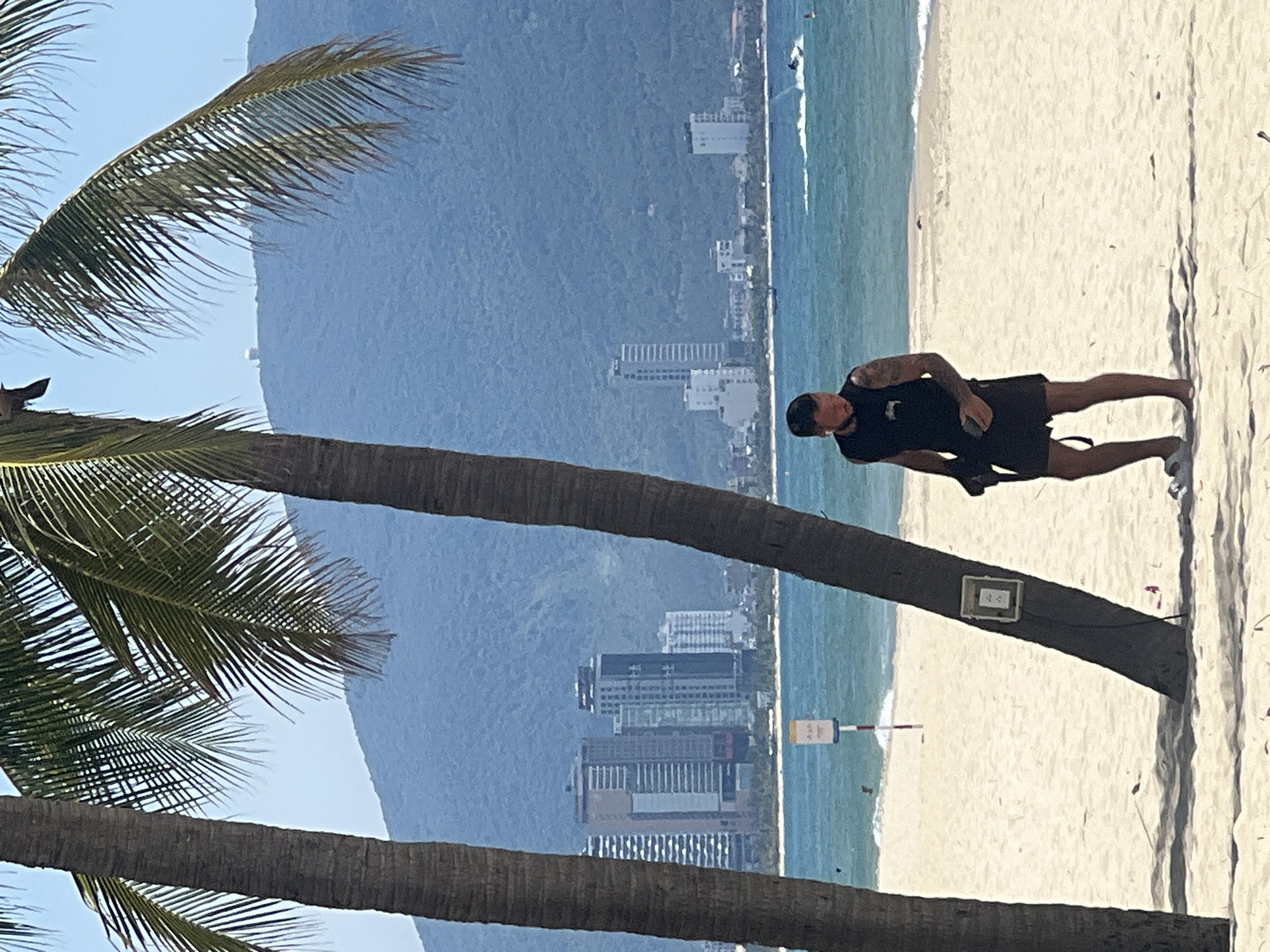 The image depicts a beach scene in Đà Nẵng, Vietnam, likely Mỹ Khê Beach, with palm trees in the foreground. A man wearing a black tank top and black shorts walks on the sand, holding a black bag. The ocean is visible with a jet ski in the background. A line of high-rise buildings forms the city skyline behind the beach, with a green, mountainous landscape rising behind the city. The sky is clear and blue.