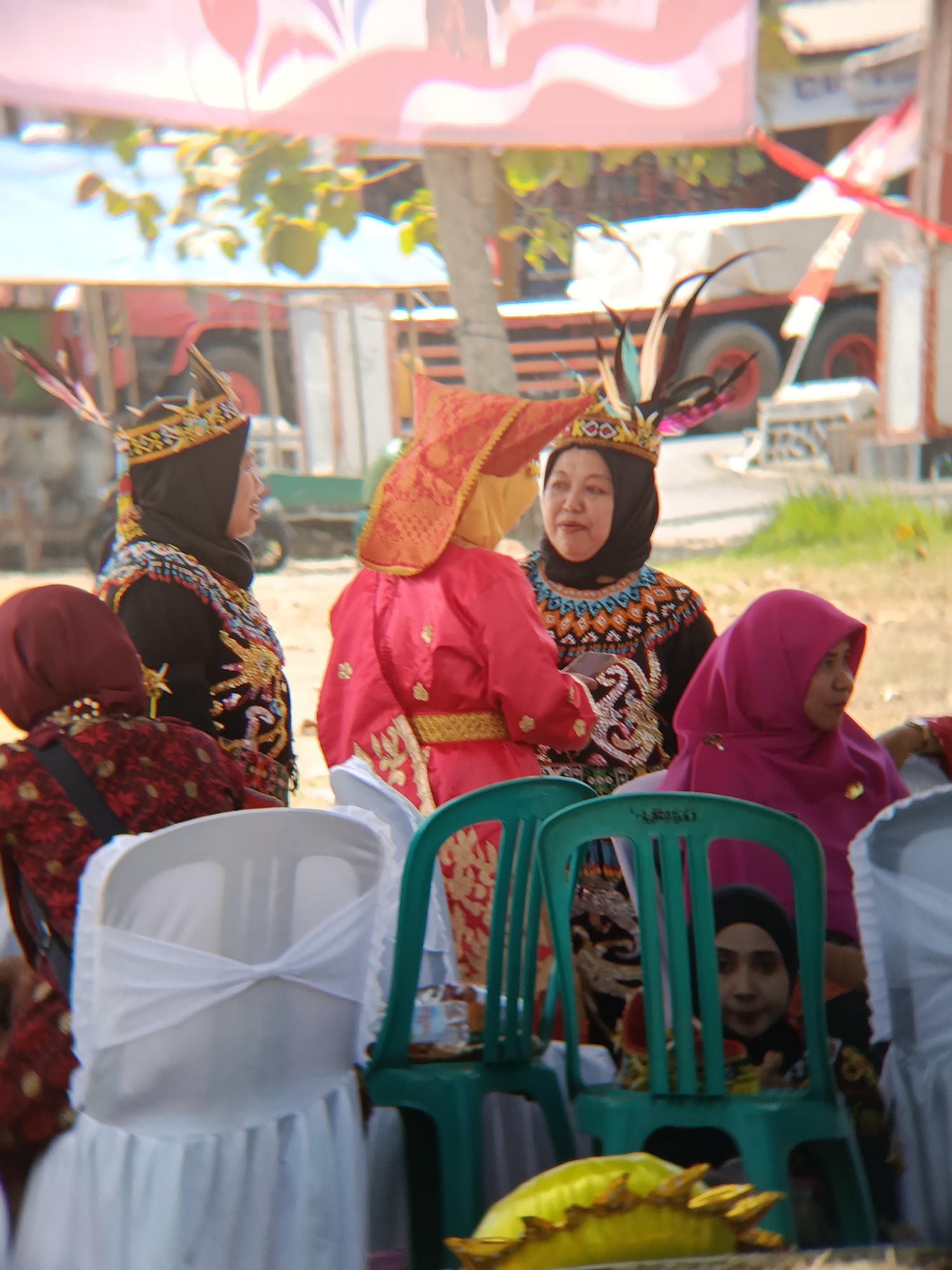 In Desa Kebon Manis, Kecamatan Cilacap Utara, Indonesia, a group of women are gathered outdoors, possibly for a cultural event or ceremony. The scene features several individuals dressed in traditional attire, including headscarves and ornate clothing with elaborate patterns. One woman in the foreground wears a black hijab and a black top adorned with colorful geometric patterns and gold embroidery, topped with a crown-like headdress featuring feathers. To her right, another woman is partially visible, wearing a vibrant pink hijab and looking to the side. Behind them, a woman in a bright red, traditional outfit with gold trim is seen from the side. In the background, a red truck with a white trailer is parked, and foliage is visible. Several chairs are present, some draped with white fabric, indicating a seating arrangement. The ground appears to be dusty or sandy. A person in a red and black patterned garment is seated in the lower left, facing away from the camera. Another person, also wearing a black hijab, is visible sitting behind a green plastic chair in the lower right. The overall atmosphere suggests an outdoor gathering with a focus on traditional dress and community.