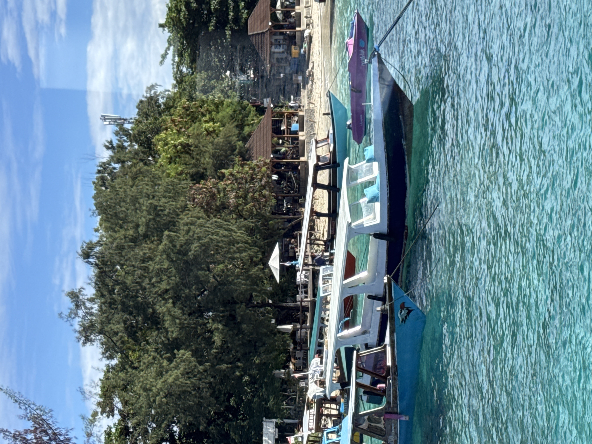 This image depicts a vibrant and serene tropical coastal scene, likely a beach in Indonesia, given the context.
The foreground is dominated by clear, shallow, turquoise-colored water, showing ripples and reflecting sunlight. Several long, narrow wooden boats, characteristic of the region (possibly jukungs or similar tour boats), are moored in the water parallel to the shore. These boats are brightly painted; one prominent boat is white and blue, another is purple, and others have white canopies or structures. On the side of the purple boat, the text "pantai lok" is visible. A blue flag or pennant on the bow of one blue boat displays a red and black emblem, possibly a stylized bird or mythical creature.
Behind the boats, a narrow strip of sandy beach transitions into a dense line of lush green tropical trees and shrubs, some resembling pines or palms, providing ample shade. Nestled among the greenery are several open-air wooden structures with brown roofs, appearing to be beachfront restaurants, cafes, or guesthouses. White umbrellas are visible near some of these establishments. A few indistinct figures can be seen sitting or standing near these structures, suggesting people are relaxing or dining.
The sky above is a clear, bright blue with scattered, wispy white clouds. In the upper-left, above the tree line, a tall, grey mast or telecommunications tower is visible in the distance. The scene is brightly illuminated, indicating a clear, sunny day, likely mid-morning or afternoon. The overall atmosphere is peaceful and picturesque, characteristic of a tropical vacation destination.