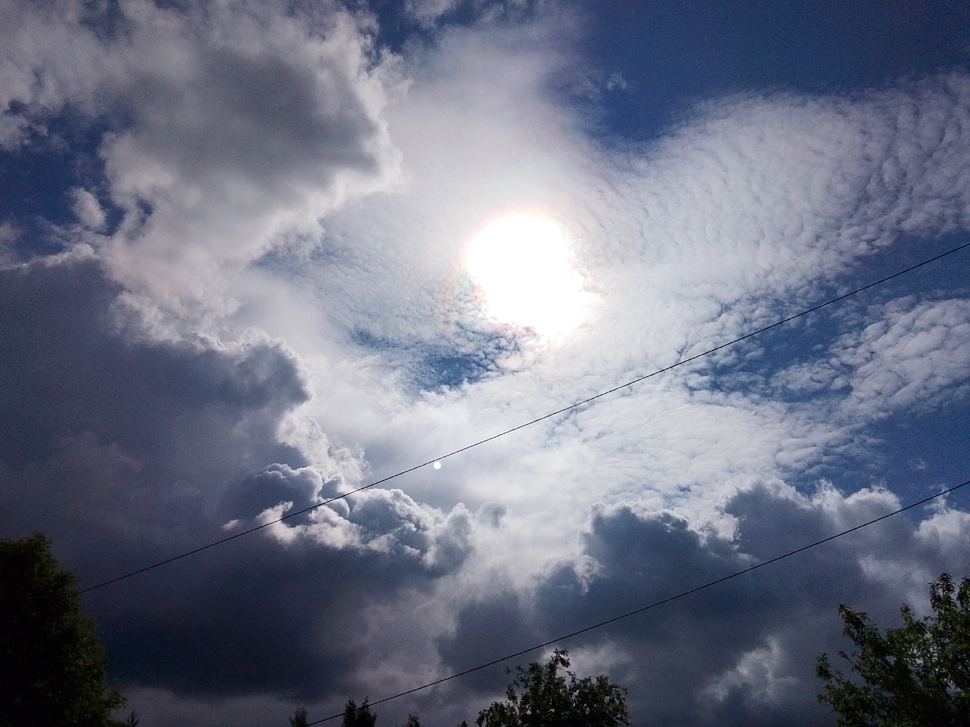 The image is a low-angle shot of a sky with cumulus and altocumulus clouds, some of which are dark and ominous. The sun is visible through a break in the clouds. There are power lines crossing the frame. In the bottom of the frame are the tops of trees. The location is likely Снопок Новый, Russia.