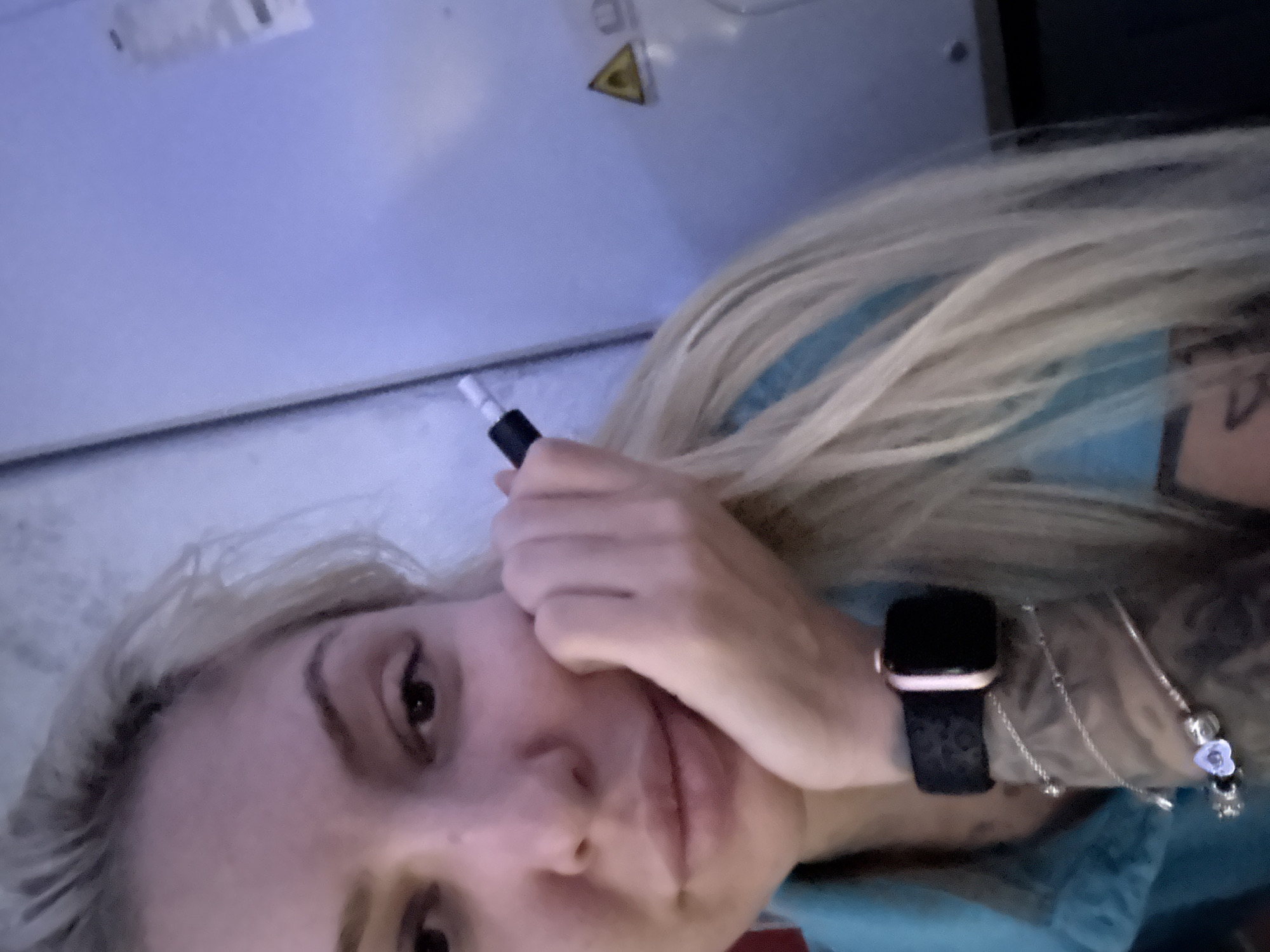 A woman with blonde hair and tattoos on her right arm is shown in a close-up, tilted selfie. She is holding a vape pen near her face. She is wearing a black Apple watch and bracelets on her left wrist. The background includes a blue-tinted wall with a yellow triangle hazard symbol on it. The location is impossible to determine.
