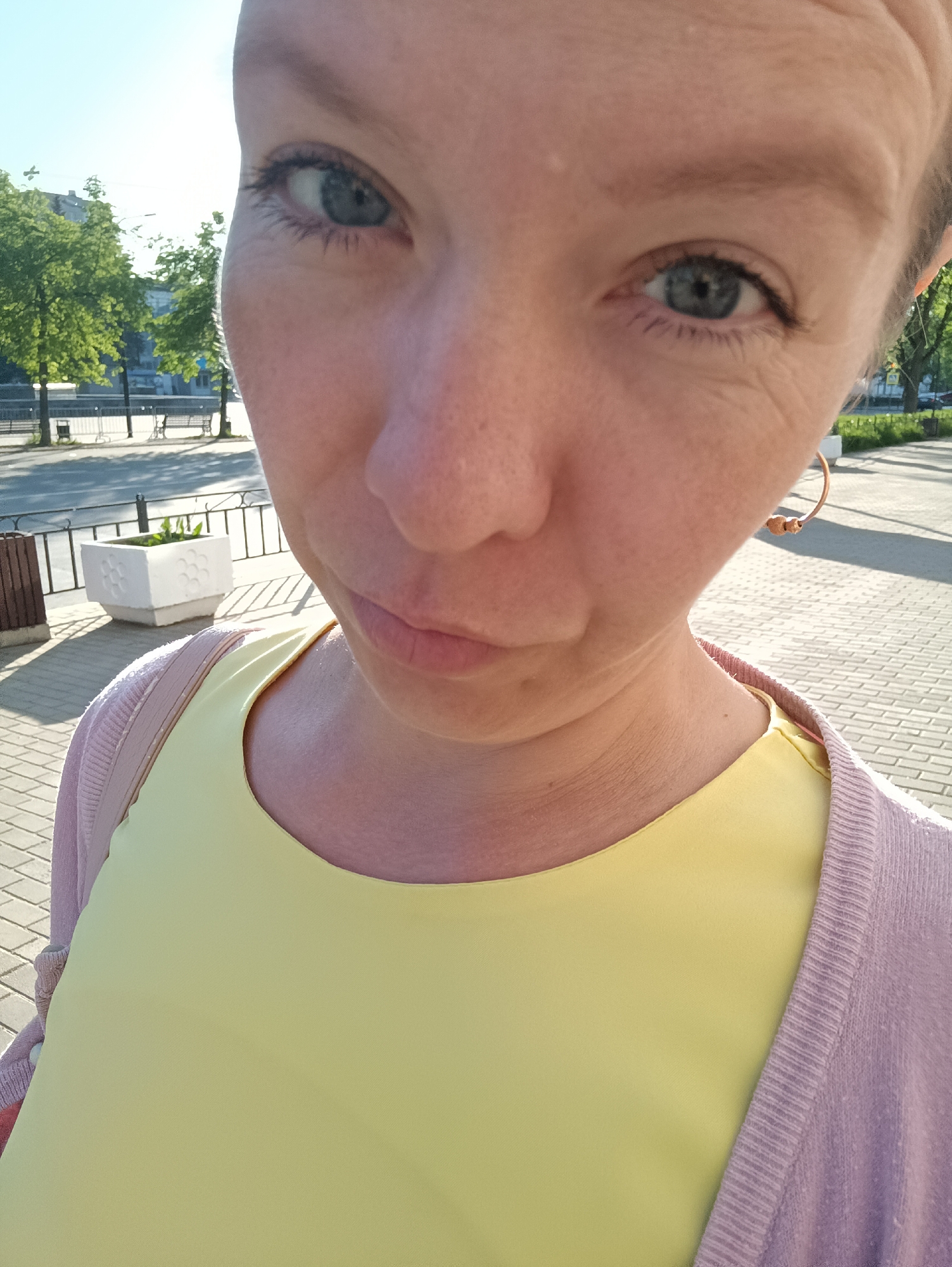 A close-up, slightly upward-angled selfie photograph shows a light-skinned person with blue eyes, wearing a yellow top and a light purple cardigan. A gold earring is visible on the person's right ear.
The background features a street scene in the city of Chaikovsky, Russia. A brick-paved walkway is visible with a white planter box. A street with a metal railing is further back, and trees are visible in the background. Buildings are also visible. The light suggests it is daytime.