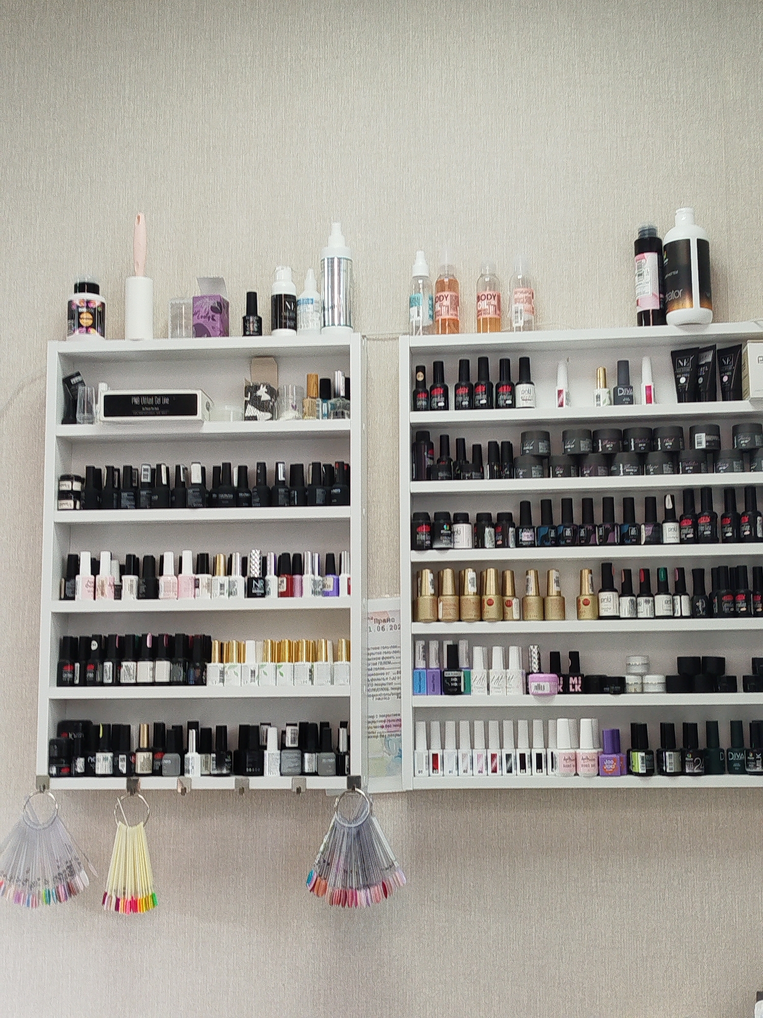 The image shows a wall-mounted display of nail polish and related products in what appears to be a nail salon. The display consists of white shelves holding numerous bottles of nail polish in various colors, as well as other beauty products such as lotions and creams. There are also display stands for nail polish colors hanging from hooks on the wall below the shelves. The shelves are organized with different types and brands of products. The background is a textured, light-colored wall. No people are visible in the image. The image is focused on the product display, with the environment being secondary. The location is Yekaterinburg, Russia, as indicated by the text on a flyer or poster partially visible on the right side of the image. The flyer contains Cyrillic text, typical of Russian signage, and a date "1.06.202" indicating a date in June.