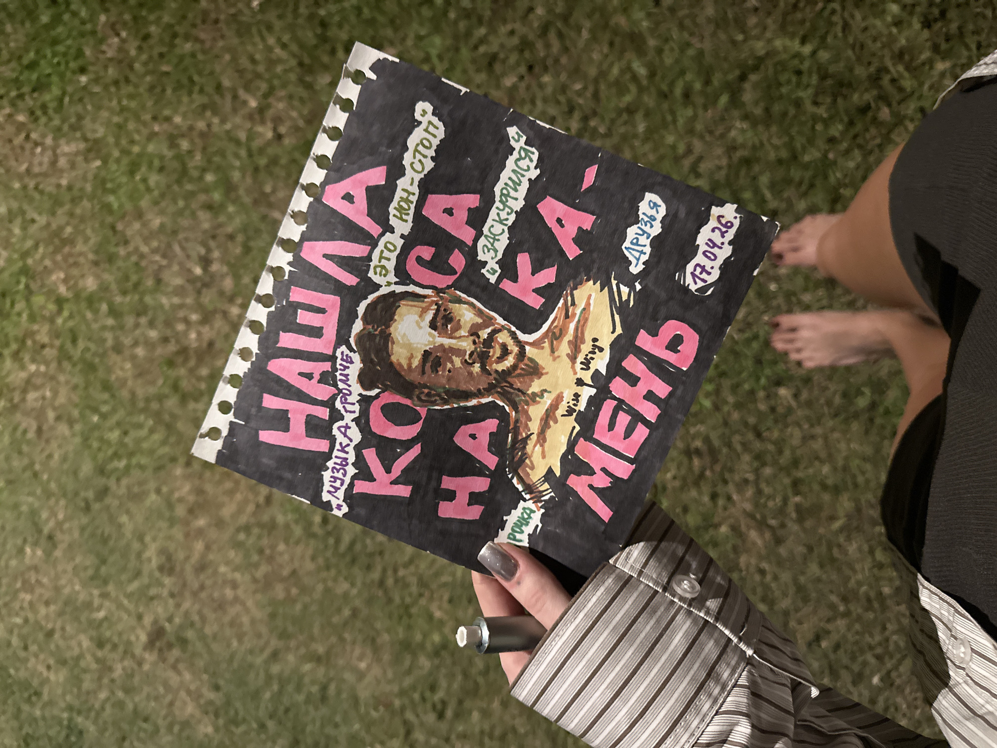 An overhead photograph shows a person holding a vibrant, hand-drawn artwork against a backdrop of green grass. The artwork, created on a piece of paper with torn, perforated edges, likely from a spiral notebook, features a dark background with bold pink and white Cyrillic text. A stylized illustration of a bearded man's face, rendered in brown and yellow tones, is central to the design.
Prominent Cyrillic text on the artwork includes "НАША КОСА" (Nasha Kosa), "ЗАСКУФИЛСЯ" (Zaskufilsya), and "МЕНЬ" (Men'). Other phrases and words written in various styles and colors are "ЭТО, НОН-СТОП" (This is non-stop), "МУЗЫКА ГРОМЧЕ" (Music Louder), "ДРУЗЬЯ" (Friends), and "РОЧКА" (Rochka). The English words "Wise Wiry" are also visible, along with the date "17.04.26."
The hand holding the artwork is adorned with gray nail polish and wears a striped long-sleeved shirt. This hand also grips a small, cylindrical metallic object, possibly a vape pen. In the blurred background, another person's bare feet and lower legs, partially covered by dark clothing, are visible on the grass. The scene takes place outdoors on a bright day, in a grassy area, consistent with the specified location of Ubud District, Indonesia. The image captures a casual moment, possibly presenting a personal artistic creation or a promotional piece.