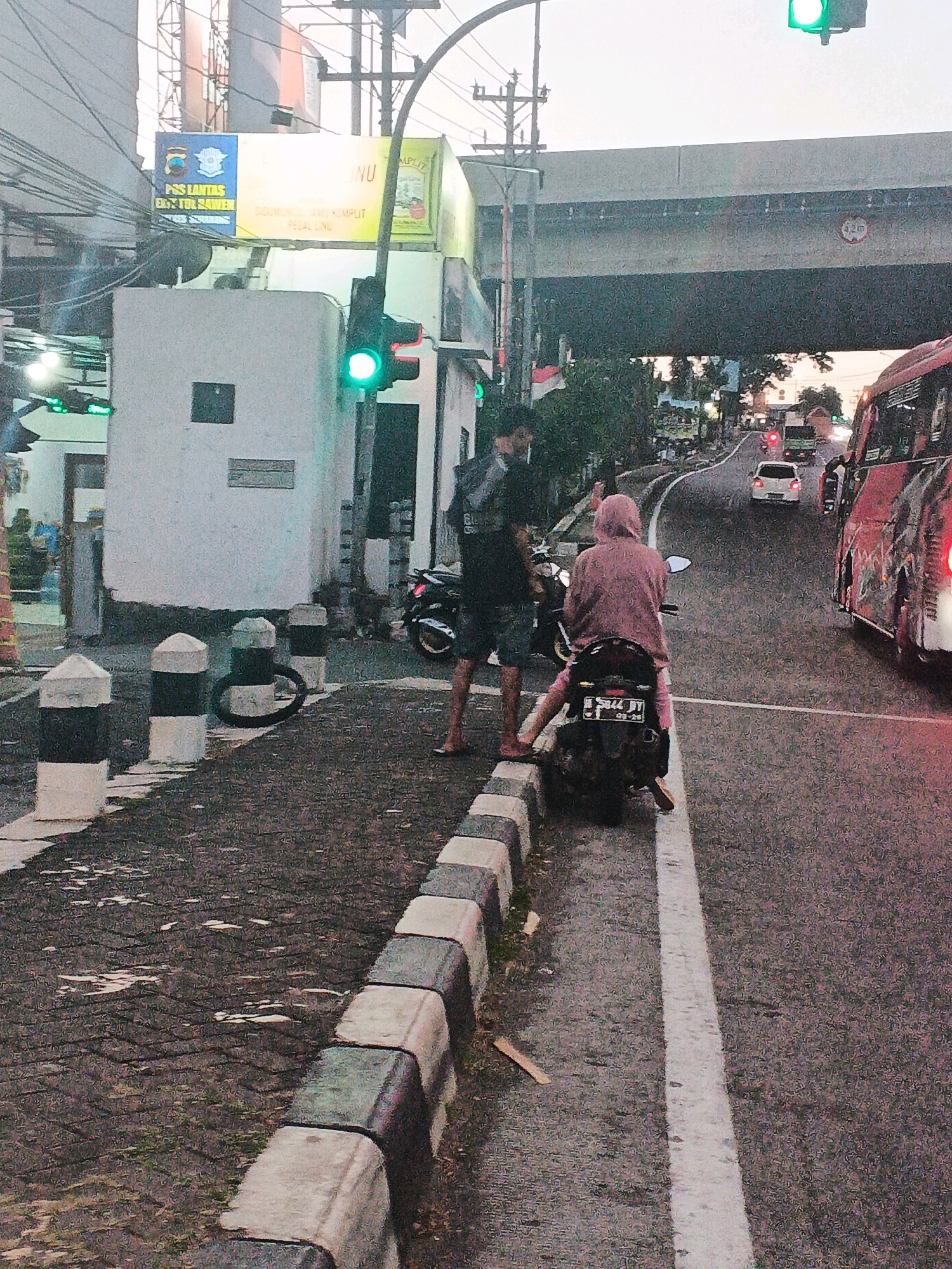 The image depicts a street scene in Tegalrejo, Indonesia, possibly at an intersection. In the foreground, a person in a pink headscarf and matching pants is seated on a black motorcycle. To their left, a person wearing a dark shirt and shorts stands. A traffic light is visible in the upper left quadrant, showing a green signal.
In the middle ground, a white building with a dark square on its facade is present. Several concrete bollards, alternating black and white, line the sidewalk. Beyond the people and the motorcycle, a paved road curves to the right.
The background features an elevated road or bridge structure, with a sign indicating a height restriction of 4.2 meters. Traffic is present on the road, including a red bus on the right side of the frame and other vehicles further down the road. The sky appears overcast or at dusk, with muted lighting. A sign with text in Indonesian and a logo is mounted on a pole above the white building. The overall scene suggests a typical urban environment with active traffic and pedestrian activity.