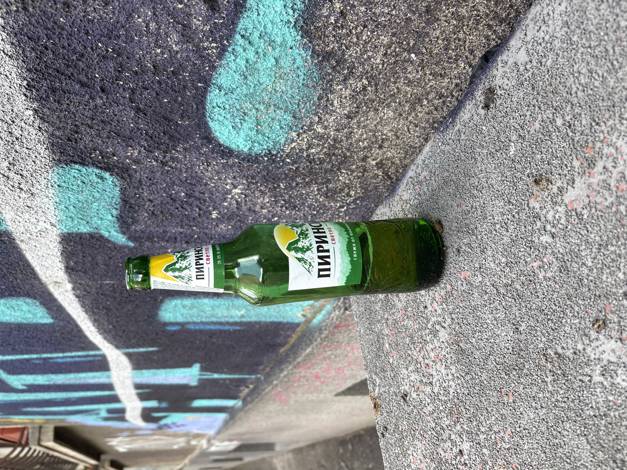 A green glass bottle lies horizontally on a coarse, speckled grey and reddish-pink concrete or granite surface. Behind the bottle is a textured wall or surface covered in graffiti, featuring broad strokes of dark purple/blue, bright turquoise/teal, and white paint. The scene is illuminated by bright daylight, highlighting the rough textures of the materials.
The bottle appears to be a standard beer or beverage bottle. Its label, primarily white and green, clearly displays text in Cyrillic script. The most prominent word is "ПИРИНСКО" (PIRINSKO), identifying a popular Bulgarian beer brand. Below this, the word "СВЕТЛО" (SVETLO), meaning "light," is visible. Near the bottle's neck, a date "29.09.26" is printed, likely indicating an expiration or best-before date. The label also features a stylized image of a mountain and some greenery, characteristic of the Pirinsko brand.
The graffiti on the background surface consists of abstract shapes and lines, suggesting an urban outdoor environment. The overall impression is one of an item discarded in a public space. No people are present, and no discernible activity is taking place. The visible Cyrillic text on the bottle strongly suggests a location within a Slavic-speaking country, specifically Bulgaria, consistent with the provided context of Sofia.