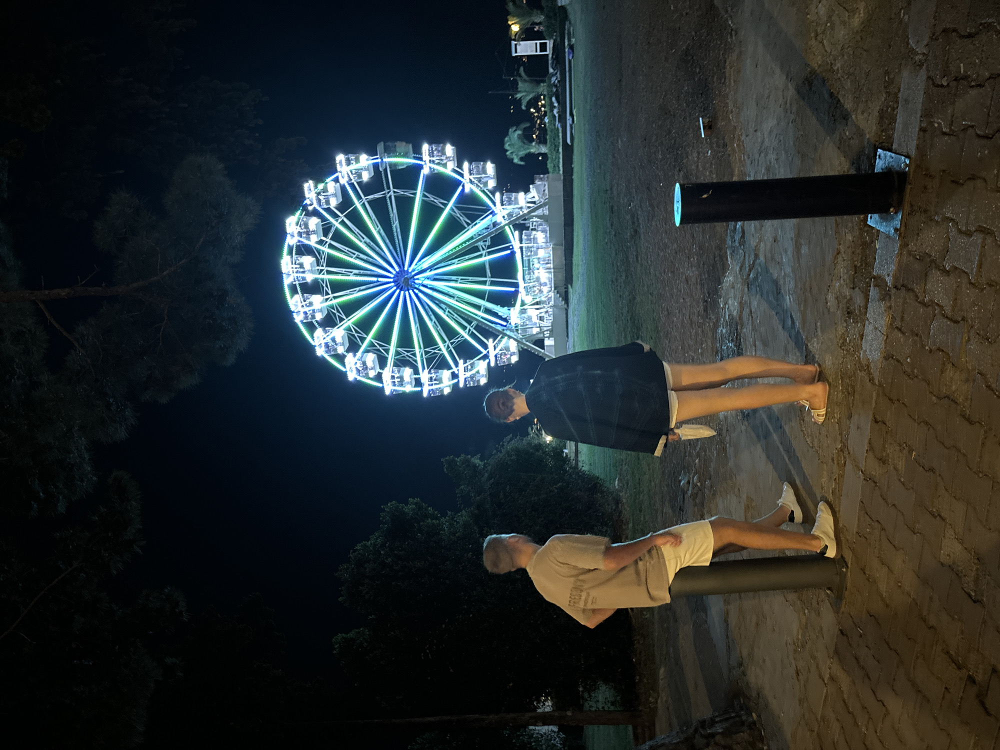 Two individuals stand on a brick pathway at night, facing a brightly lit Ferris wheel in Budva, Montenegro. The Ferris wheel is illuminated with a vibrant blue and green light display. The individual on the left is a male wearing a tan t-shirt and white shorts. The individual on the right is a female wearing a dark jacket, white shorts, and sandals. They are standing near a dark, cylindrical bollard. The background consists of a grassy area, trees, and distant lights, suggesting an amusement park or public space. The ground is paved with interlocking bricks and some areas appear damp.