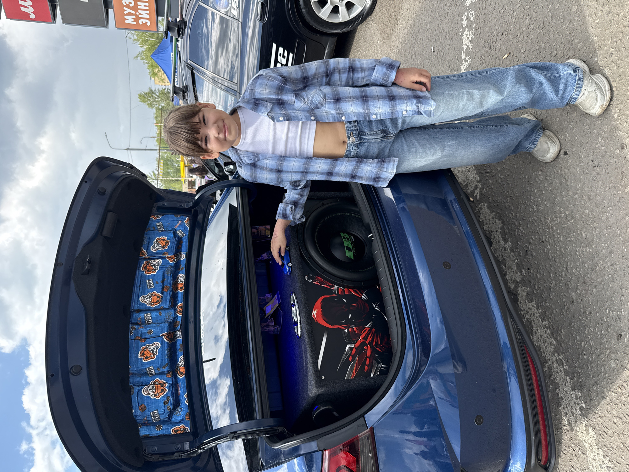 A medium shot depicts a young person reclining on the open trunk of a blue car, within an urban setting.
The person, of indeterminate gender, is casually dressed in a white undershirt, open blue plaid shirt, and light-wash jeans. Their posture is relaxed, leaning on the trunk.
The trunk interior is custom-fitted with audio equipment, including a large subwoofer and other components illuminated with blue LED lighting. A panel within the trunk features an illustration of a red spider-like character. The underside of the trunk lid is lined with a blue fabric featuring a pattern of stylized orange and white figures.
The car is parked on an asphalt surface. In the background, another vehicle is partially visible, along with urban infrastructure, including signage with Russian characters. The sky is overcast. The location appears to be a parking area within the city.