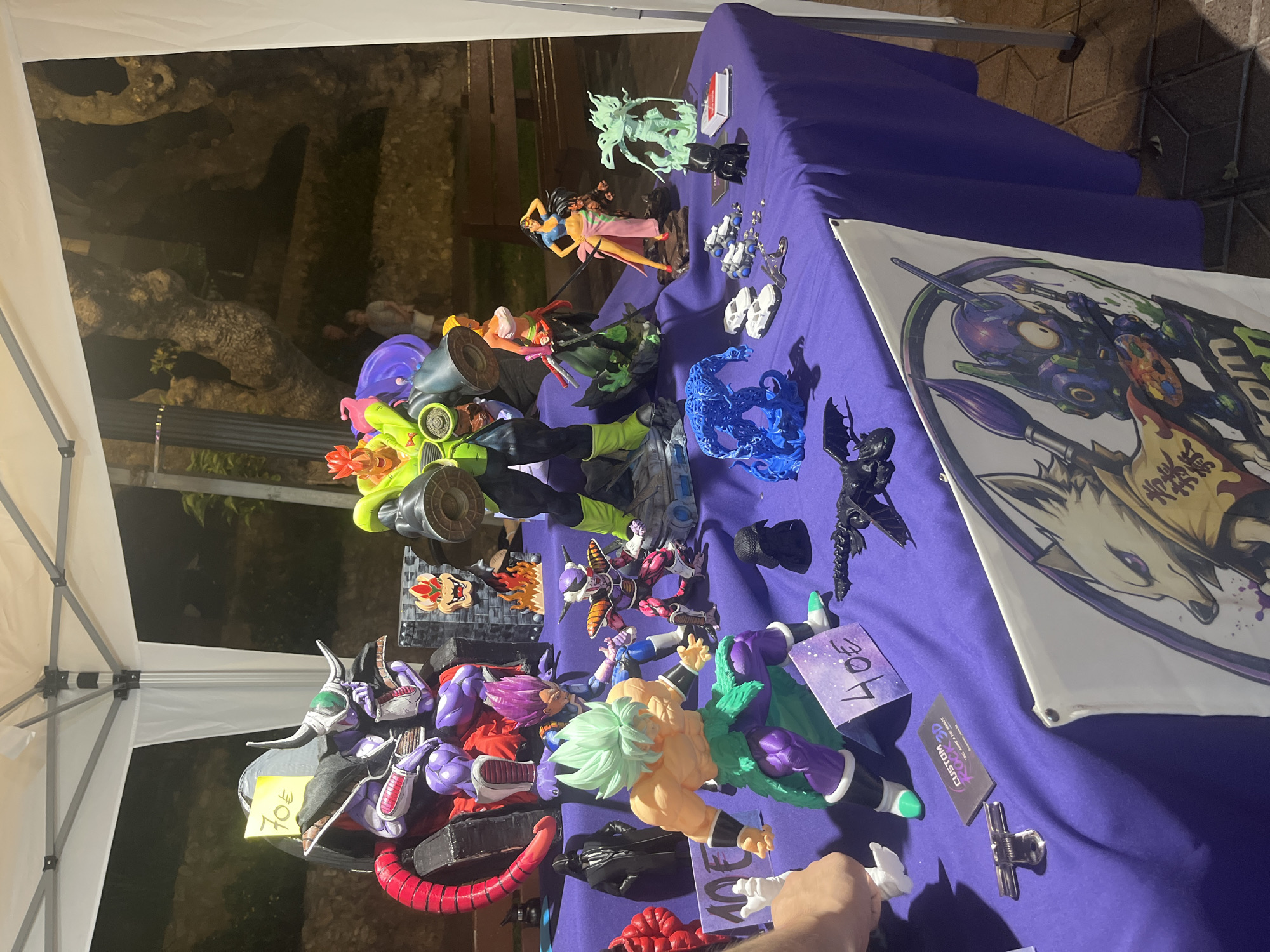 An outdoor stall is set up under a white tent with a purple tablecloth covering a table laden with anime action figures and merchandise. The stall is likely in an outdoor market or event space in Tarragona, Spain, given the "CUSTOM RO3D" business card visible, which appears to be a local 3D printing service.

The figures displayed include characters from Dragon Ball Z, such as Broly in his Super Saiyan form, Frieza, and Vegeta, as well as a figure that resembles Cell. There are also figures of characters that appear to be from other anime series, possibly including a female character with a pink skirt and a character with a green suit and large goggles. Other items on display include miniature cars, a black dragon figure, a blue, flame-like creature, and a black figure that looks like Darth Vader. Several price tags are visible, with one indicating "70€" and another "40€". A banner with illustrations of animals and a robot holding paintbrushes is hanging from the front of the table. Two people are partially visible in the background, standing near trees. The lighting suggests it is evening or night.