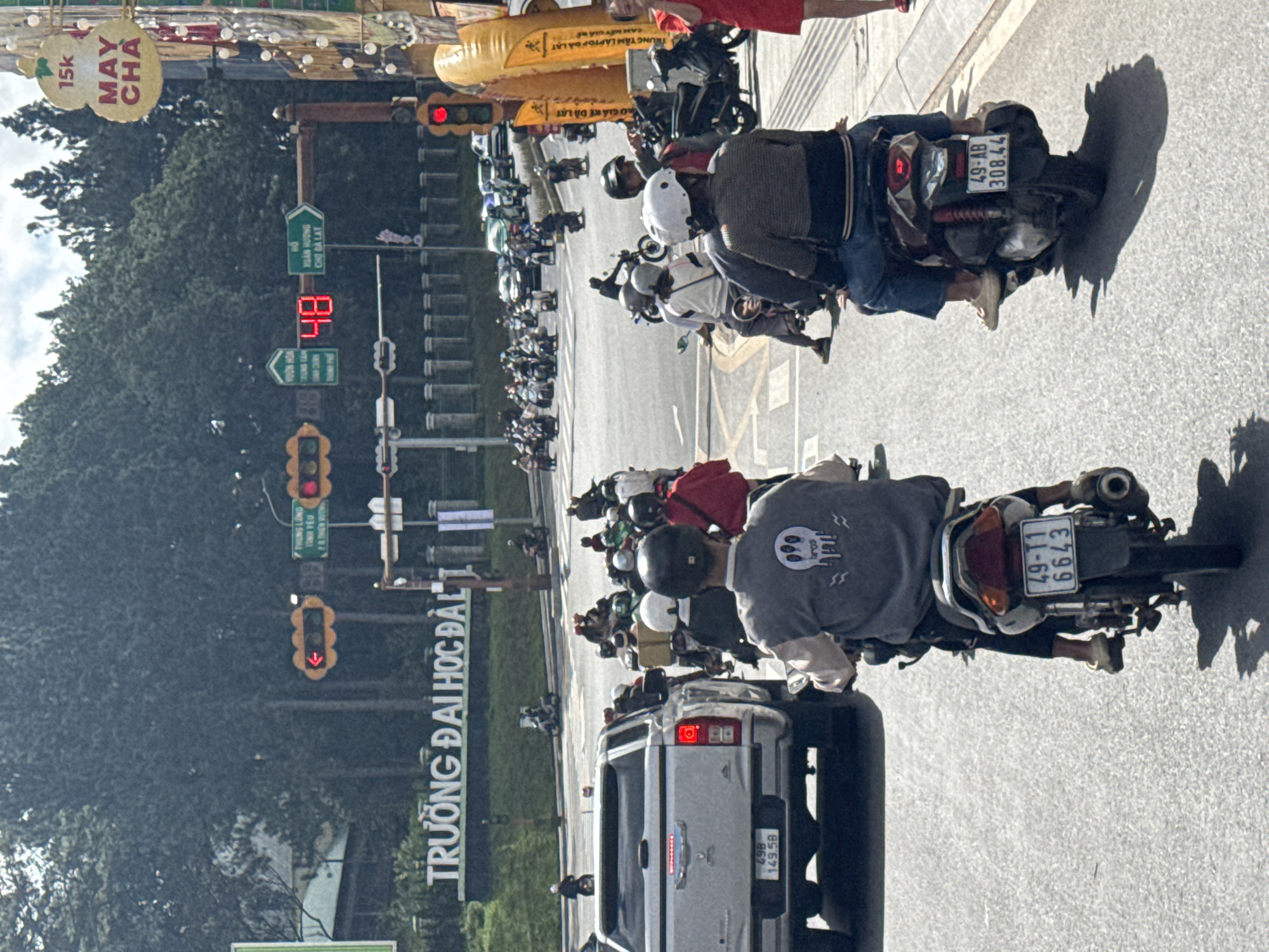 The image depicts a busy intersection in Da Lat, Vietnam, with numerous motorcycles and a few cars navigating the traffic. A large sign prominently displays "TRUONG ĐẠI HỌC ĐÀL" which translates to "ĐÀ LẠT UNIVERSITY". Traffic lights are visible, with one displaying a red arrow and another showing the number "48" in digital format, presumably indicating a countdown or a specific traffic signal status. Several individuals are seen riding motorcycles, some wearing helmets and casual clothing. A silver pickup truck is stopped at the intersection. On the right side of the image, a tall yellow inflatable advertising structure is present, along with a sign that reads "MAY CHA". The environment appears to be an urban street scene on a bright, possibly sunny day, with lush green trees forming a backdrop. The ground is paved asphalt.