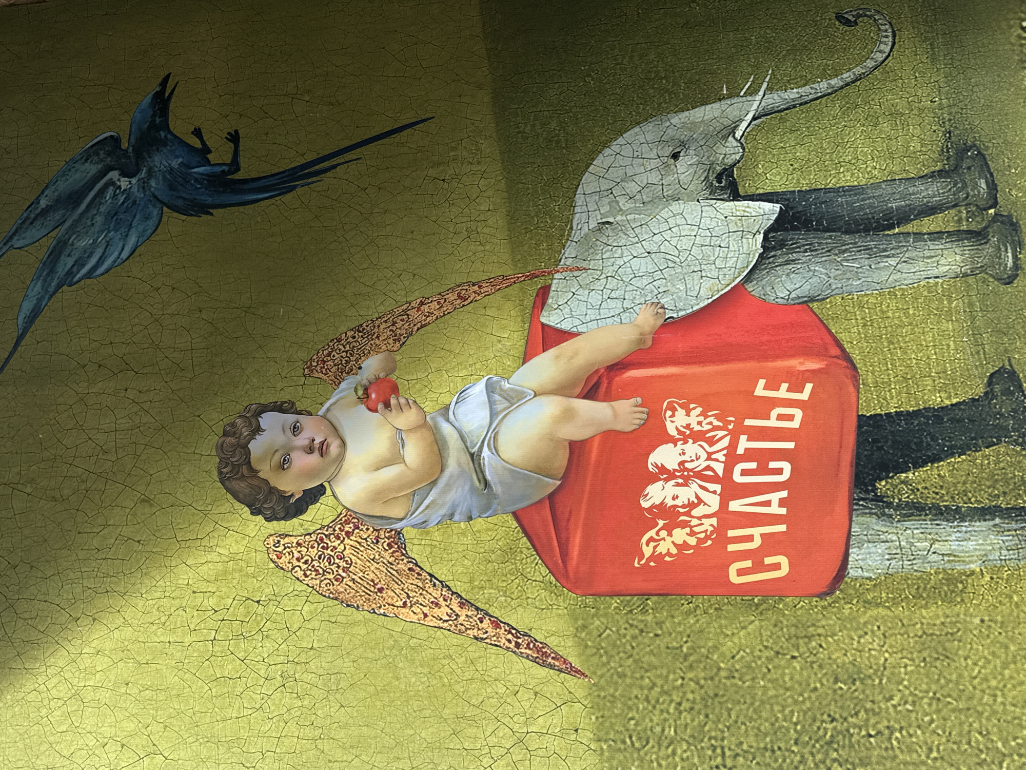 The image depicts a surreal tableau on a surface with a cracked, aged appearance. A dark blue bird with outstretched wings is positioned in the upper left corner. An infant angel, holding a small red apple, reclines on a red rectangular object. The object is labeled "СЧАСТЬЕ" (Happiness in Russian). An elephant with cracked skin stands behind the red rectangle. The elephant wears trousers and shoes. The scene is set against an ochre-colored backdrop. This is a detail of a painted advertising sign from the Eliseev Emporium in Saint Petersburg, Russia.