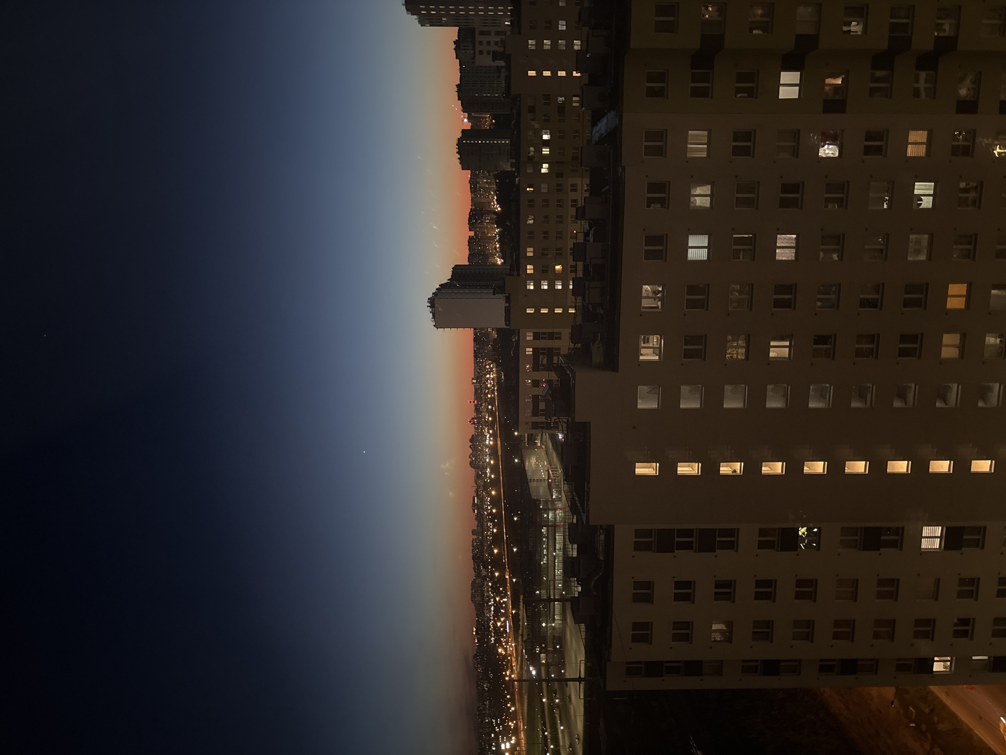 The image depicts a nighttime cityscape in Vsevolozhsk, Russia. A tall apartment building, densely populated with lit windows, dominates the right side of the frame. To the left, the city extends into the distance, with streetlights illuminating roads and other buildings. The sky transitions from dark blue to a lighter orange/red hue near the horizon, suggesting either sunset or sunrise. There are no people visible in the image, but the lit windows imply occupancy. The image conveys a sense of urban density and the transition between day and night.