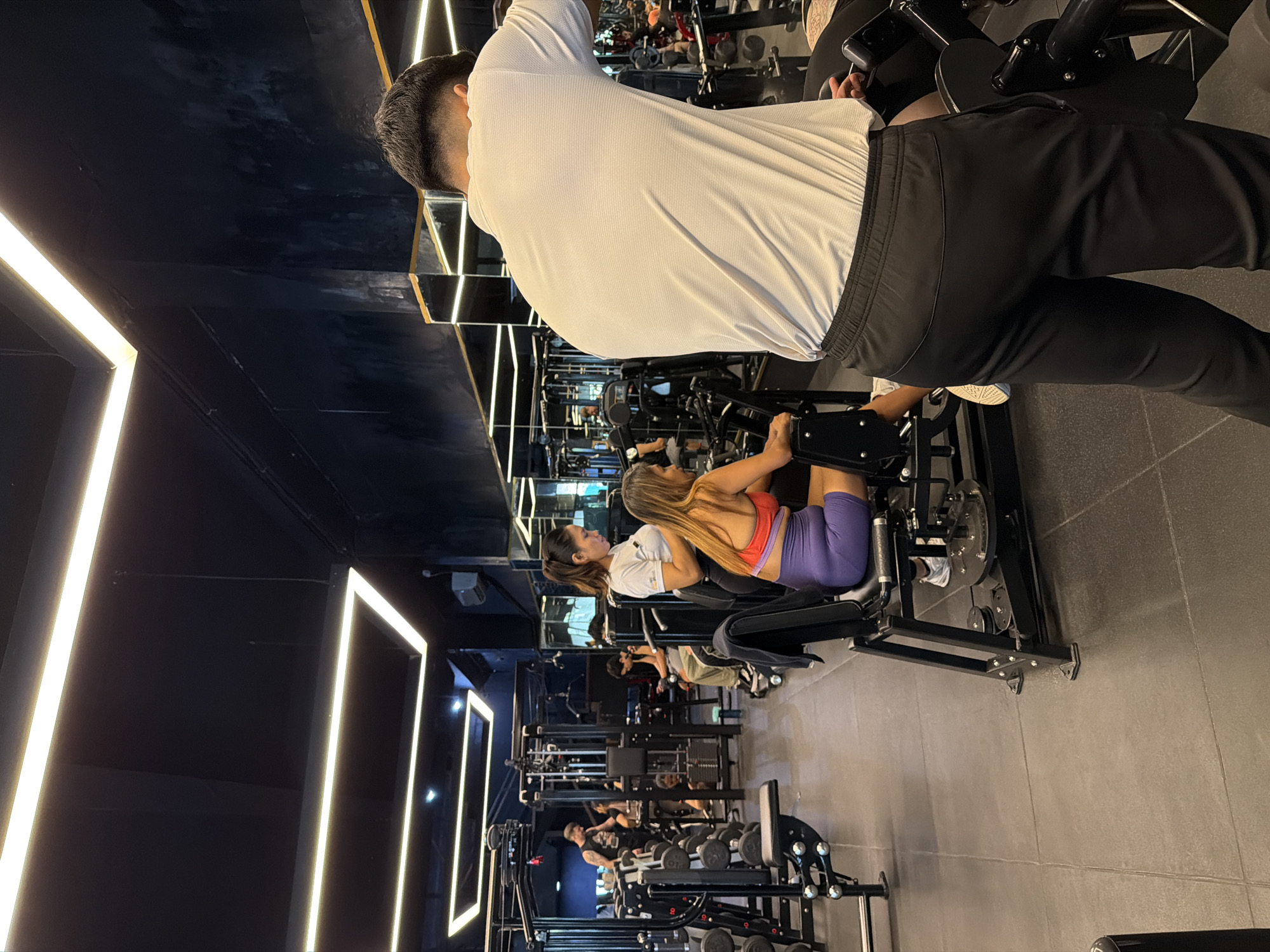 Inside a gymnasium in Denpasar, Indonesia, a medium shot depicts gym-goers using various weight machines. A woman with long brown hair wears a white t-shirt, a red sports bra, and purple shorts while using a leg press machine. Behind her, an unidentified individual stands with their back to the camera, wearing a white t-shirt and black pants. Additional gym patrons are scattered throughout the facility, using other workout equipment. Fluorescent lighting accents the gym's decor.