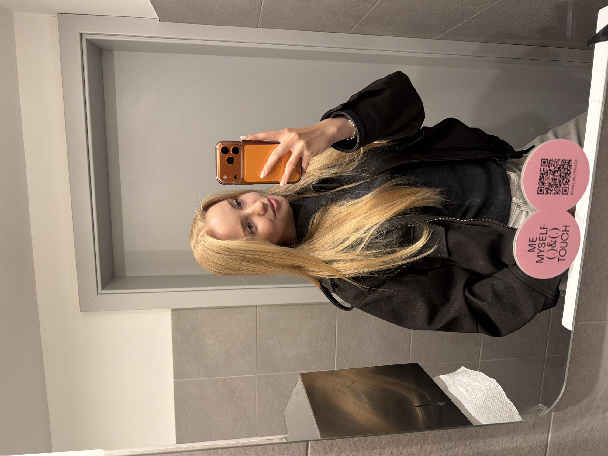 Here is a detailed description of the image:
A fair-skinned woman with long blonde hair is taking a selfie in a bathroom mirror. She is wearing a black turtleneck sweater under a black jacket. The phone she's holding has an orange case.
The bathroom has gray walls and a gray-framed mirror. In the bottom of the mirror's reflection, there is a metal paper towel dispenser with paper towels sticking out of it. A pink sticker with the text "ME MYSELF (.)&(.) TOUCH" and a QR code is attached to the right edge of the mirror.