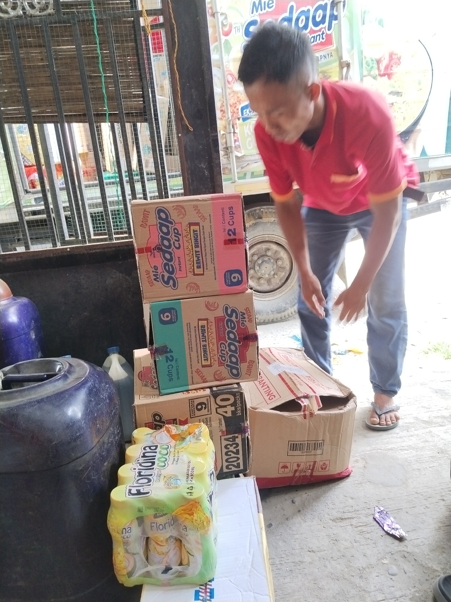 A man in a red polo shirt and blue jeans is bending down in front of a stack of boxes. He is in an outdoor setting with a truck in the background. There are several stacks of boxes and containers visible. In the foreground, there is a package of "Floridina Coco Orange" drinks. To the left, there are two large blue plastic containers. To the right of the man, a partially opened cardboard box is visible. The scene appears to be in a commercial or distribution area, possibly a street or loading dock, in Sawit Seberang, Indonesia. The overall atmosphere is casual and functional.