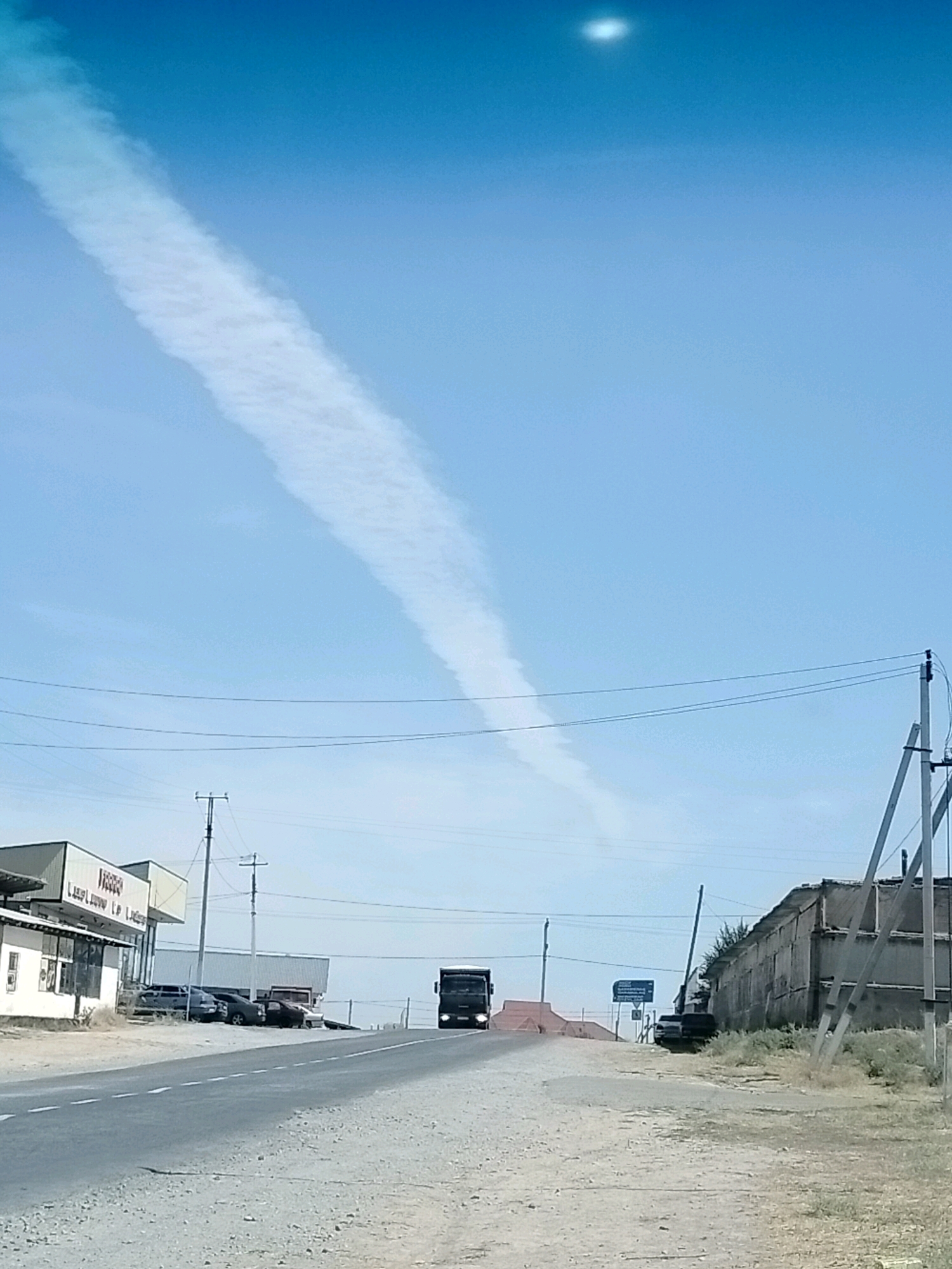 A wide-angle, eye-level outdoor shot depicts a road in a semi-arid environment, likely the city of Kölkent, Kazakhstan, under a clear blue sky. A long, thin contrail stretches diagonally across the upper portion of the sky, originating from the top left and extending towards the center. A faint, bright circular object, possibly the sun or a lens flare, is visible near the top center of the sky. The road, paved with asphalt, curves slightly and ascends towards the right side of the frame. On the left side of the road, a low, rectangular building with a white facade and a grey awning is visible. Several cars are parked in front of it. Across the road, to the right, are several power poles with overhead wires. Further down the road, a dark-colored truck is approaching from the distance. On the right side of the road, a long, low, dilapidated building with a corrugated metal roof and grey walls is situated. The ground is covered with gravel and dry vegetation.