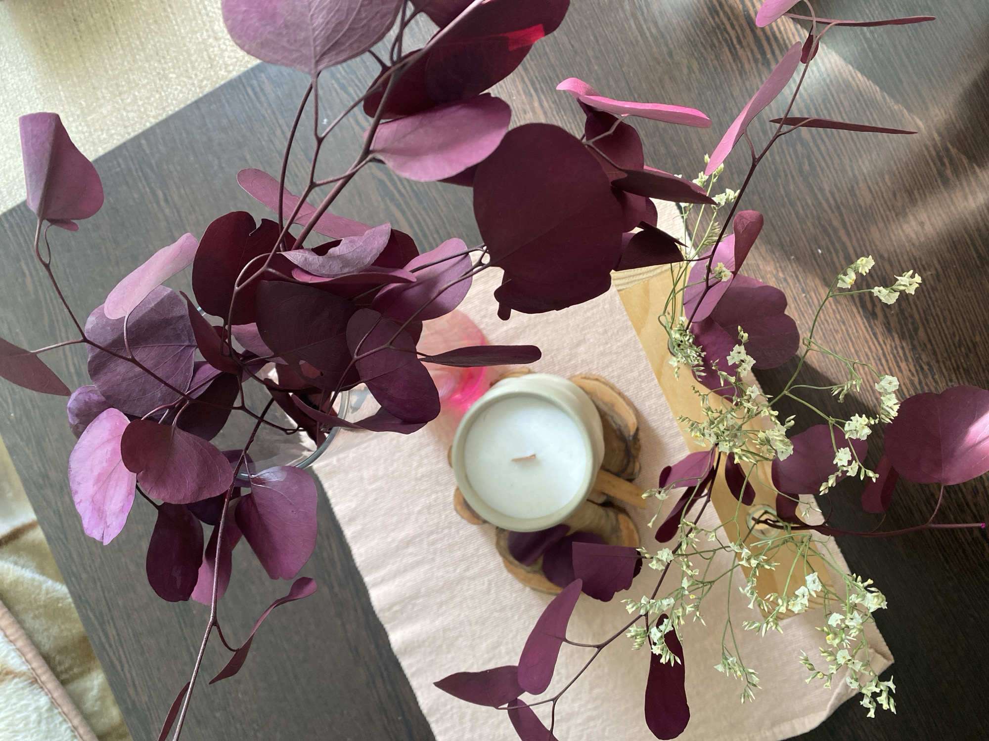 A high-angle, close-up shot shows a tabletop display. A white candle in a light-colored ceramic holder sits on a small wood slice stand on a light pink table runner. Purple leaves and sprigs of small white flowers are arranged around the candle. The table is dark brown. The photograph may have been taken in Chaikovsky, Russia, but there is no forensic evidence to support this information.