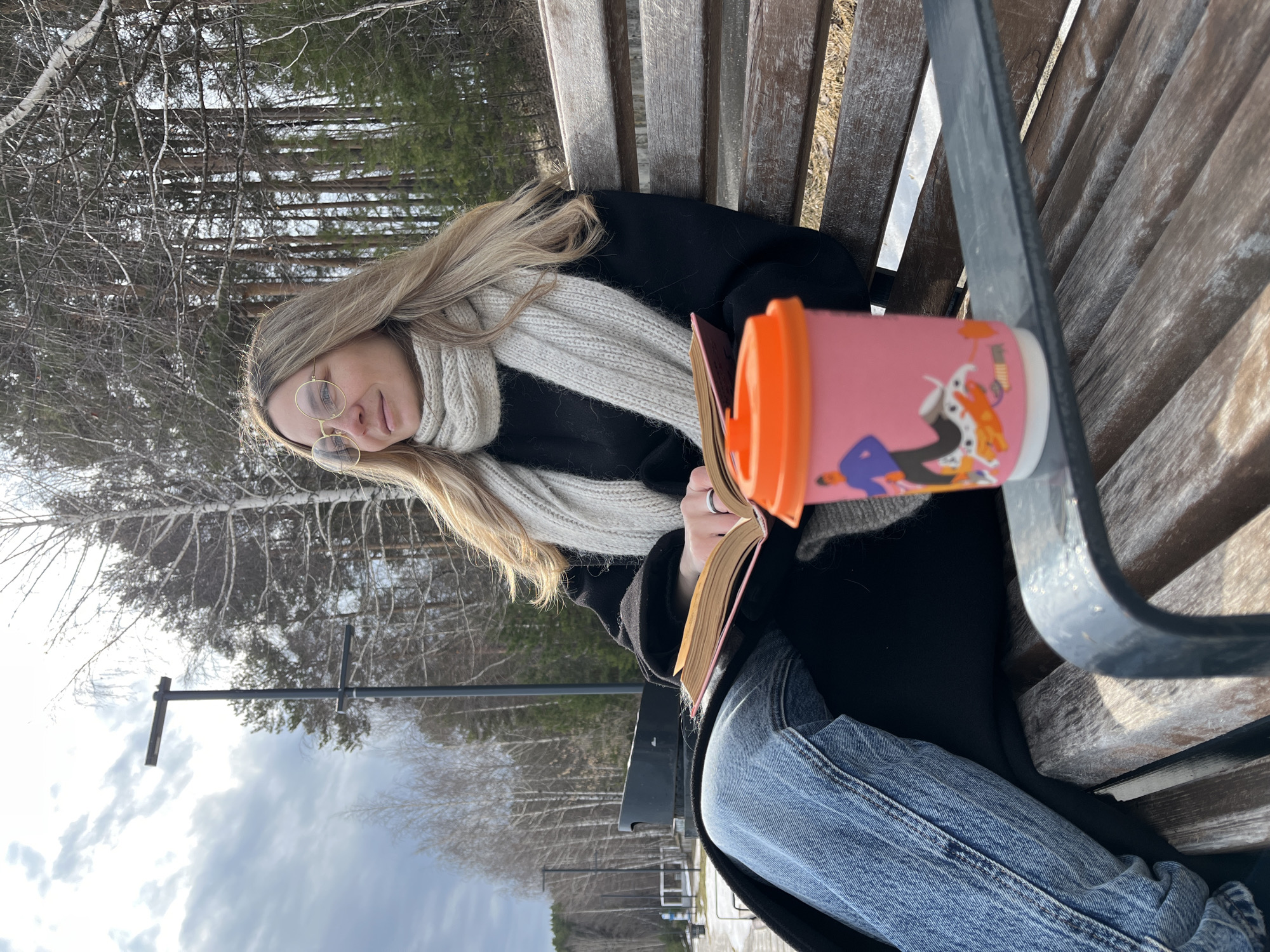 A woman is seated on a wooden park bench, reading a book. She has long blonde hair and wears round glasses, a black coat, and a light-colored scarf. A pink cup with an orange lid sits on the bench beside her. She is wearing blue jeans. The background shows trees with bare branches, indicating a colder season, and a cloudy sky. A street lamp is visible in the background. The location is outdoors, in a park setting, in Chaikovsky, Russia.