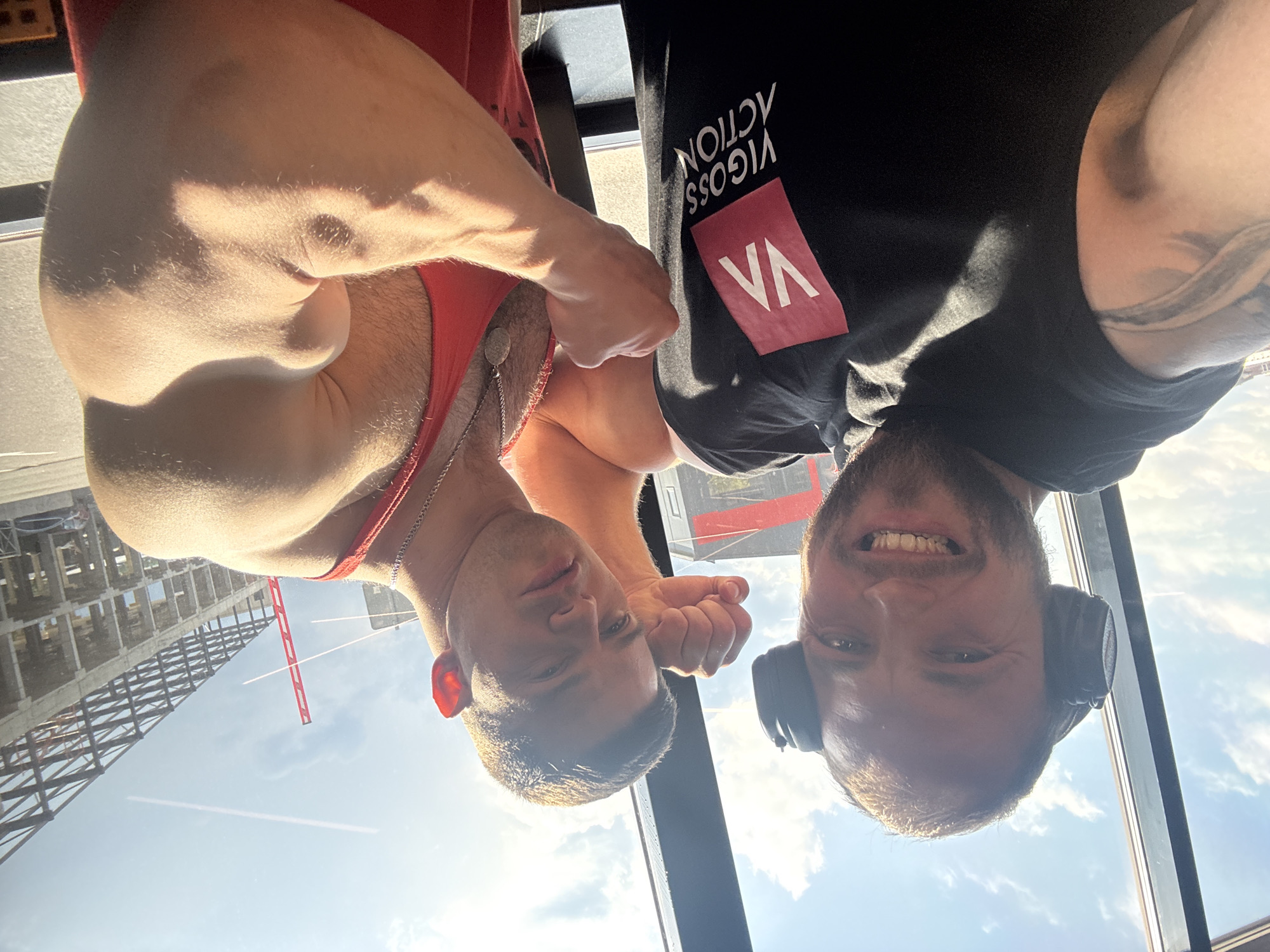 Two men are posing for a selfie in front of a window with a city view. The man on the left is wearing headphones and a black t-shirt with a red logo that says "VIGOSS ACTION". He has a beard and is smiling. The man on the right is wearing a red tank top and has a muscular arm. He is clenching his fist. In the background, there is a construction site with a large building under construction and a red crane. The sky is blue with some clouds.