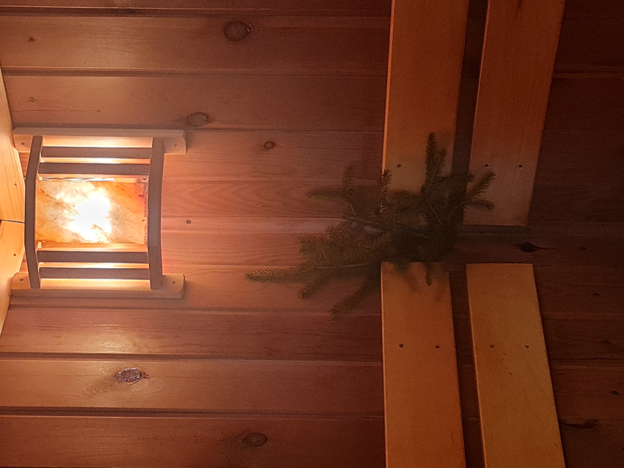 The image shows the interior of a wooden sauna with a Himalayan salt lamp. The walls are made of vertical wooden planks, and two wooden benches are visible, one above the other. A small sprig of evergreen branches is placed between the benches. The salt lamp, mounted on the wall, emits a warm, orange light. The overall scene suggests a relaxing and therapeutic environment. It is not possible to determine the location in Angarsk, Russia based on the visual information alone.