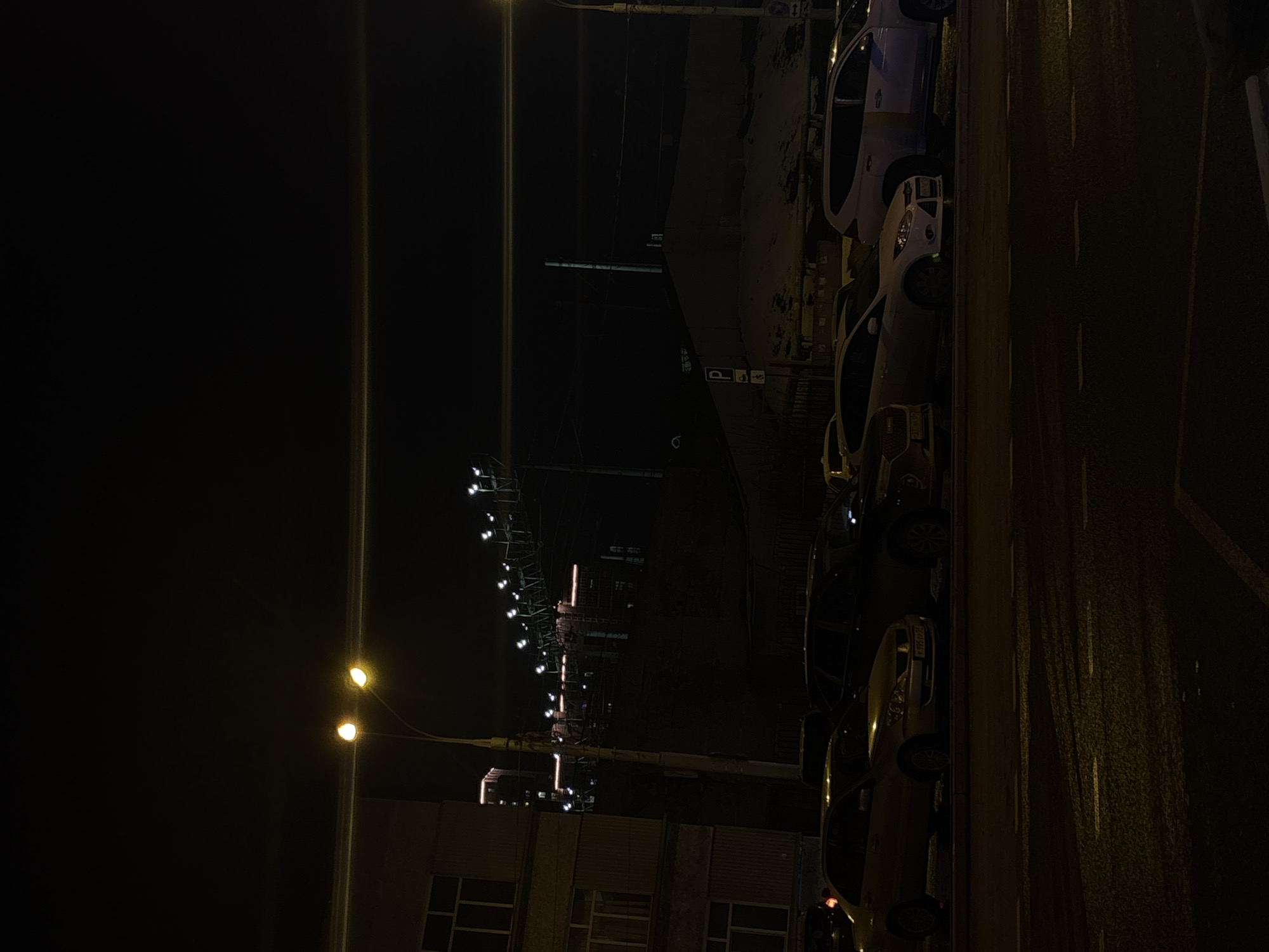 The image depicts a nighttime urban scene in Perm, Russia. A row of parked cars lines the right side of what appears to be a street. Streetlights illuminate the scene, casting a warm glow on the cars and the adjacent road. A building stands on the left, with visible windows and possibly exterior lighting fixtures. Overhead, a structure resembling a pedestrian bridge or overpass is adorned with decorative lights. The general atmosphere is dark, with only the artificial lights providing illumination.