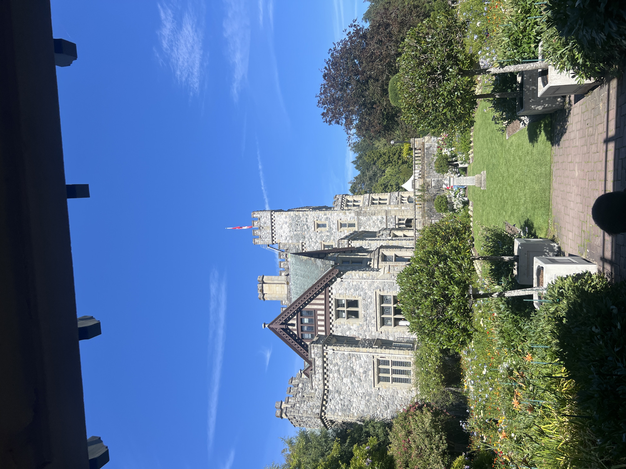 The image depicts a sunlit view of a stone castle, likely Hatley Castle, in Langford, Canada. The castle has multiple turrets and a prominent flag atop its highest tower, flying a red and white flag, possibly the Canadian flag. In the foreground, a meticulously maintained garden is visible, featuring a variety of blooming flowers in shades of orange, yellow, white, and blue, alongside manicured green shrubs and trees. A brick pathway leads towards a grassy lawn with a stone monument. The sky above is a clear, bright blue with a few wispy white clouds. A dark wooden structure, possibly part of a gazebo or pergola, frames the top of the image. The scene appears to be a tranquil, picturesque setting, typical of a historical estate or park.