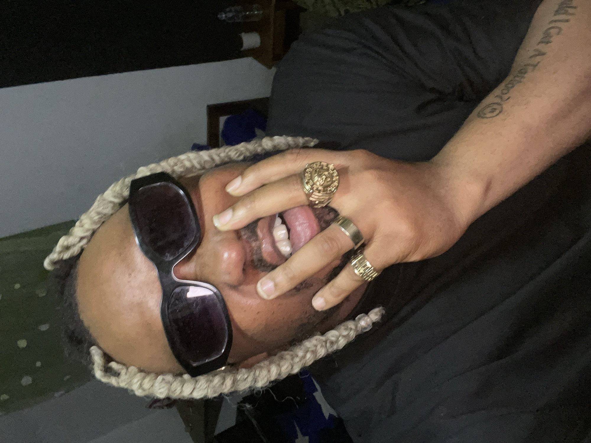 The media file captures a person, likely male, in a close-up shot, posing in an indoor setting under low light. The person's head is tilted, and they are wearing dark sunglasses. They have light-colored braided hair or dreadlocks framing their face.
The person's left hand is covering part of their mouth, with a glimpse of their tongue visible, suggesting a playful or expressive moment. Three gold-colored rings are worn on the left hand: a large, intricate ring on the ring finger, a plain band on the middle finger, and a patterned ring on the pinky finger. The person is wearing a black shirt.
A prominent detail is a tattoo on the person's right forearm, which reads "I Got A Tattoo" followed by a circular symbol. The background is dark and indistinct, with subtle hints of a wall or furniture, characteristic of an evening or night setting indoors. The image conveys a casual and expressive mood.