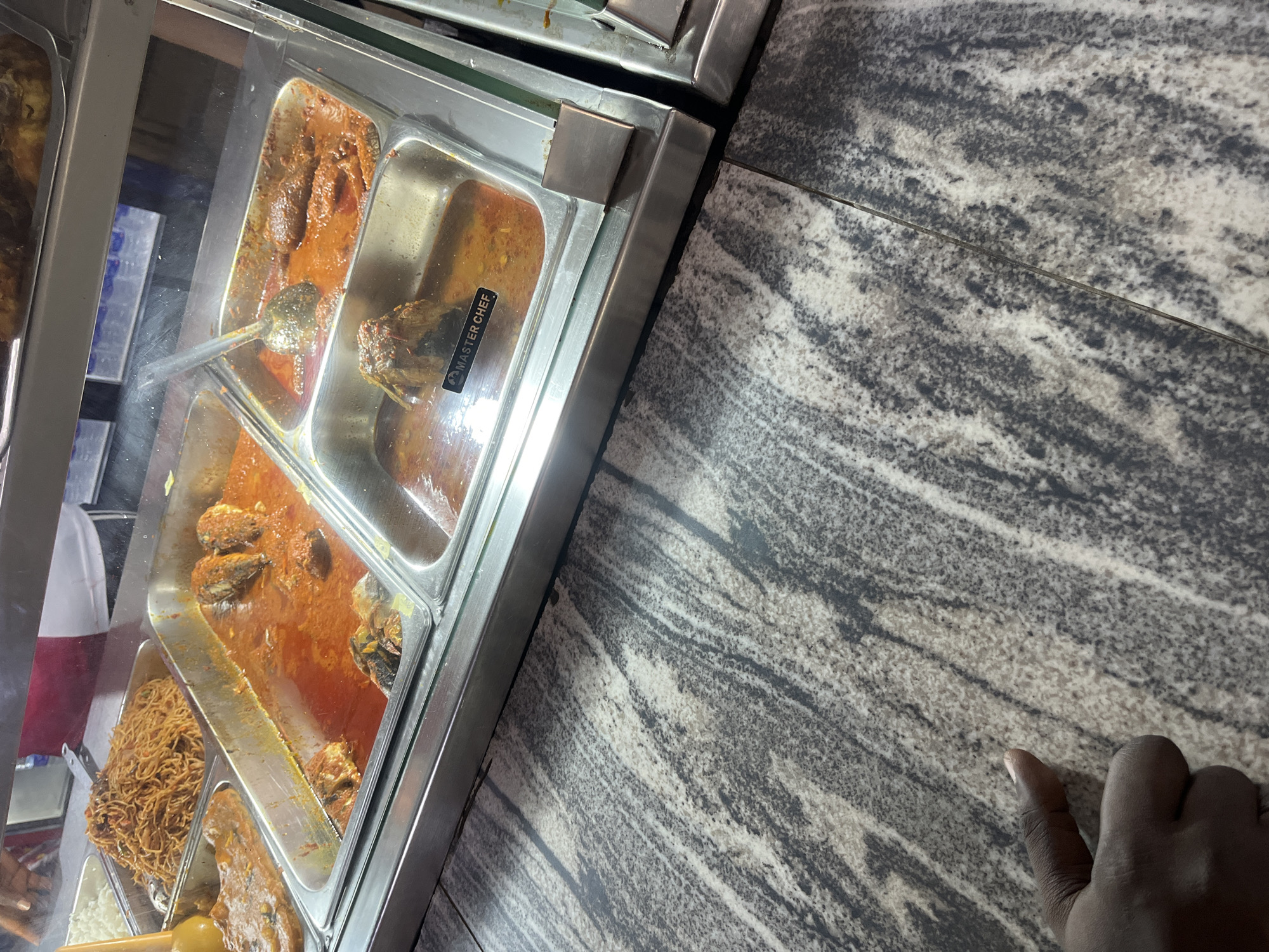 The image provides a tilted, close-up view of a food display, likely within a cafeteria or buffet setting in Osogbo, Nigeria. The scene features multiple stainless steel warming pans, commonly known as chafing dishes, filled with various dishes and covered by a clear glass shield.
In the foreground and middle, large pans hold a rich, reddish-orange stew or sauce containing sizable pieces of fish, indicating a fish stew, a common dish in Nigerian cuisine. A serving ladle is partially submerged in one of these pans. To the lower left, another pan contains a noodle dish that resembles Jollof spaghetti, characterized by its reddish-brown color and individual strands. Below it, a pan appears to hold white rice. Another pan on the far left shows a darker, perhaps meat-based, stew.
A notable detail is a black label with the text "MASTER CHEF" visible on what appears to be a food item or packaging within one of the fish stew pans.
The food display is set upon a patterned surface, possibly a granite countertop or tiled table, featuring a mix of grey, white, and dark striations. In the lower right corner, a hand with a dark skin tone is visible, resting on or near this surface, suggesting a person interacting with the food display, either waiting to be served or about to serve themselves. The lighting indicates an indoor setting, likely with artificial illumination highlighting the food. The overall scene depicts a typical ready-to-serve food counter.