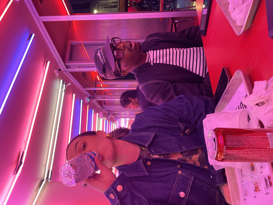 The image depicts an indoor dining scene in a modern fast-casual restaurant, located in Paris, France. The environment is characterized by an array of linear neon lights overhead, emitting strong pink, red, blue, and white illumination that reflects off glass partitions.
In the foreground, two individuals are seated at a vibrant red table. On the left, a person with dark hair, wearing a dark denim jacket over a camouflage-patterned shirt, is actively drinking from a clear plastic water bottle. This individual has visible tattoos on the left side of their neck and wears gold hoop earrings. On the right, a second person with dark skin, wearing a dark jacket over a black-and-white striped shirt, black-rimmed glasses, and a dark cap emblazoned with "L'ART DE L'AUTOMOBILE" and a car graphic, gazes towards the right with their mouth slightly open.
On the table between them, an open red Dr Pepper can with French text on its label rests on a metal tray lined with white paper, surrounded by crumpled white napkins. Another black smartphone is partially visible on the table near the right-seated individual. In the background, a third individual with dreadlocks is partially visible, seated and facing away from the camera. Several small, white security cameras are mounted on the ceiling.