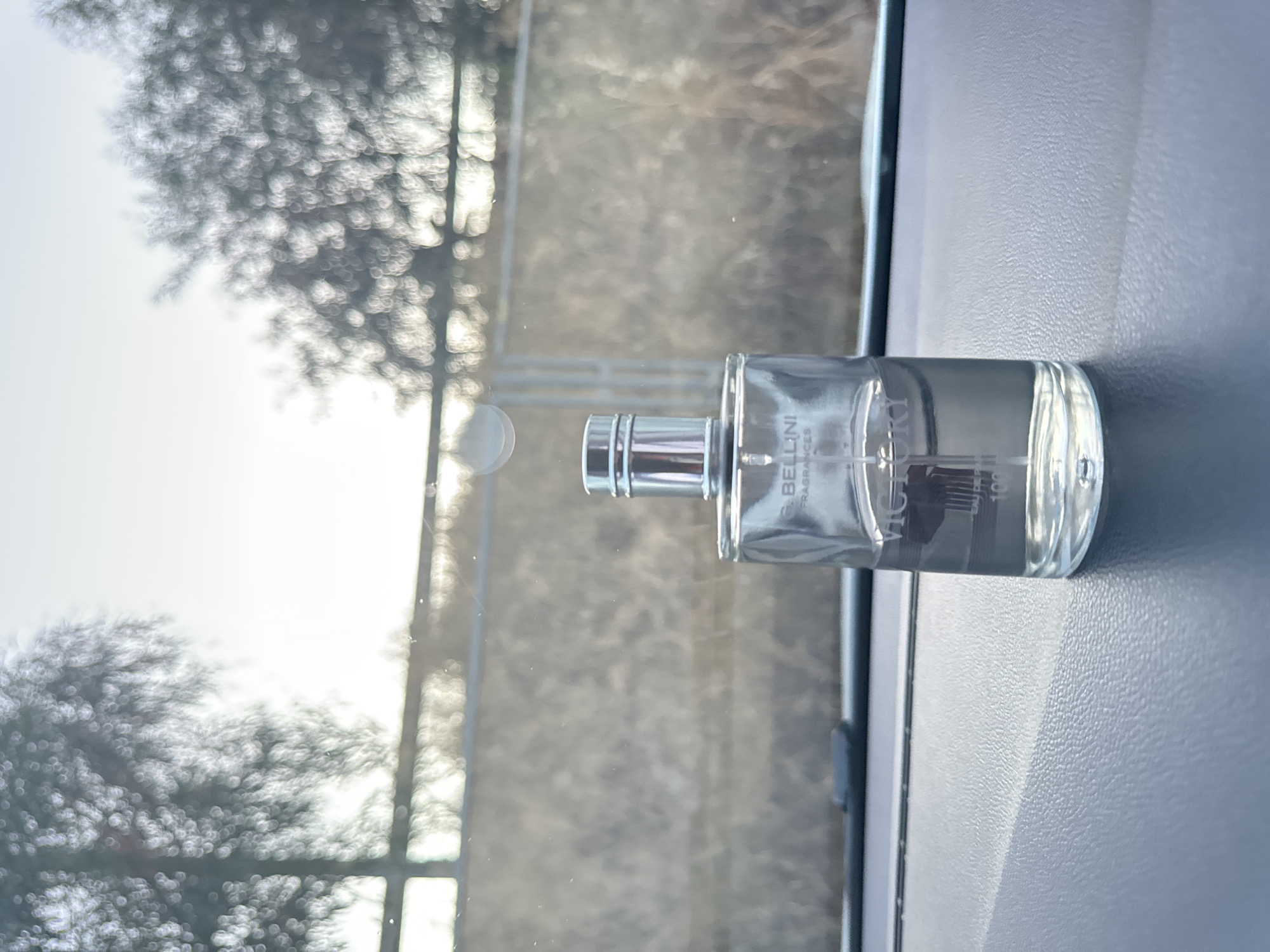 A clear glass perfume bottle with a silver cap sits on a dark gray dashboard. The bottle is partially filled with a clear liquid and has the word "VICTORY" printed on it in dark gray text. The background is blurred, showing trees and a fence, suggesting an outdoor setting. The overall lighting indicates it is daytime, possibly with the sun behind the camera creating some lens flare. There is no indication of the city of Murcia, Spain in this image.