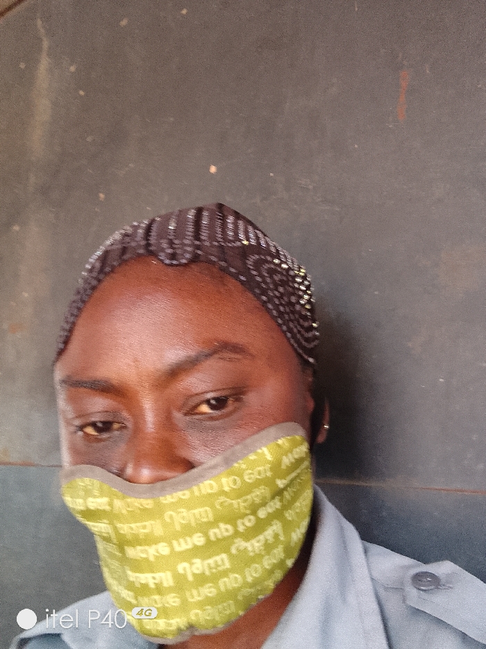 A close-up, slightly low-angle photograph depicts an individual in Hoche, Nigeria, against a plain, dark grey or dark brown textured wall. The subject has dark skin and is wearing a dark, sheer head covering adorned with reflective beads or sequins in linear and circular patterns. A light olive-green fabric face mask covers the lower half of the face, from below the eyes to the chin. The mask features repeating, inverted white or light-colored text, including the phrase "to eat," and has a lighter-colored trim. The individual's eyes are visible, looking slightly downwards, and their dark eyebrows are well-defined. A portion of a light grey or light blue-grey collared shirt with a visible buttoned shoulder tab is seen on the subject's right. A "itel P40 4G" watermark is present in the lower left corner of the image.