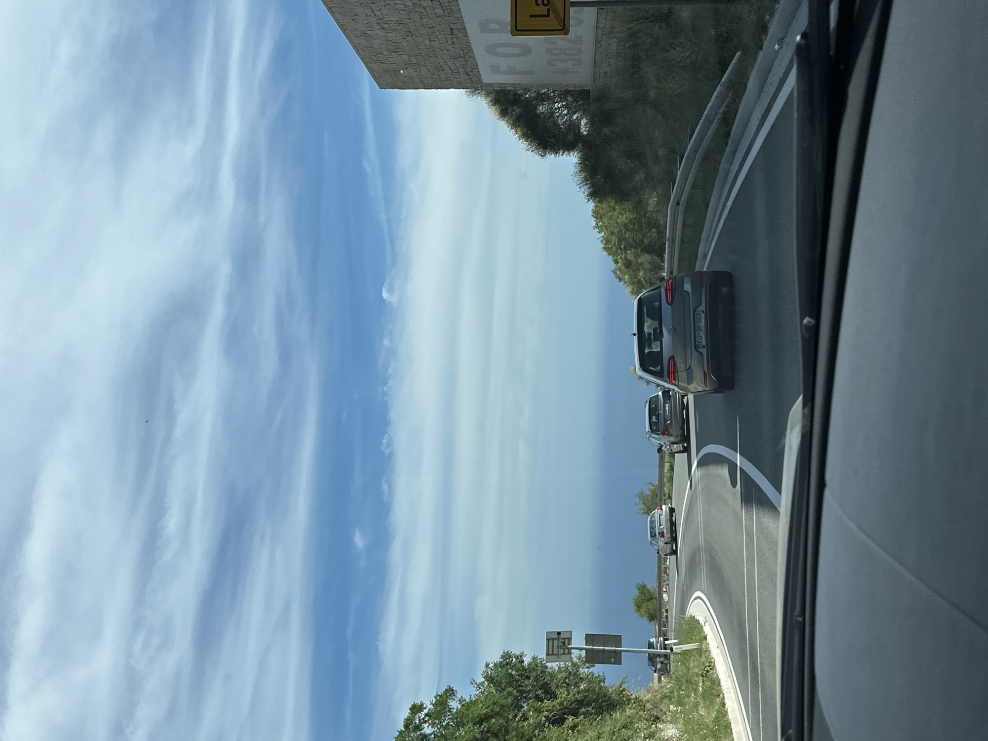 The image captures a view from inside a vehicle driving on a road in Vrela, Montenegro. The road curves to the left, with a steep, vegetated embankment on the left and a stone-faced building with a large "FOR" sign on the right. Several cars are visible on the road ahead. The sky is a bright blue with wispy white clouds, and the sea is visible in the distance. The overall scene suggests a sunny day with moderate traffic on a scenic coastal road.