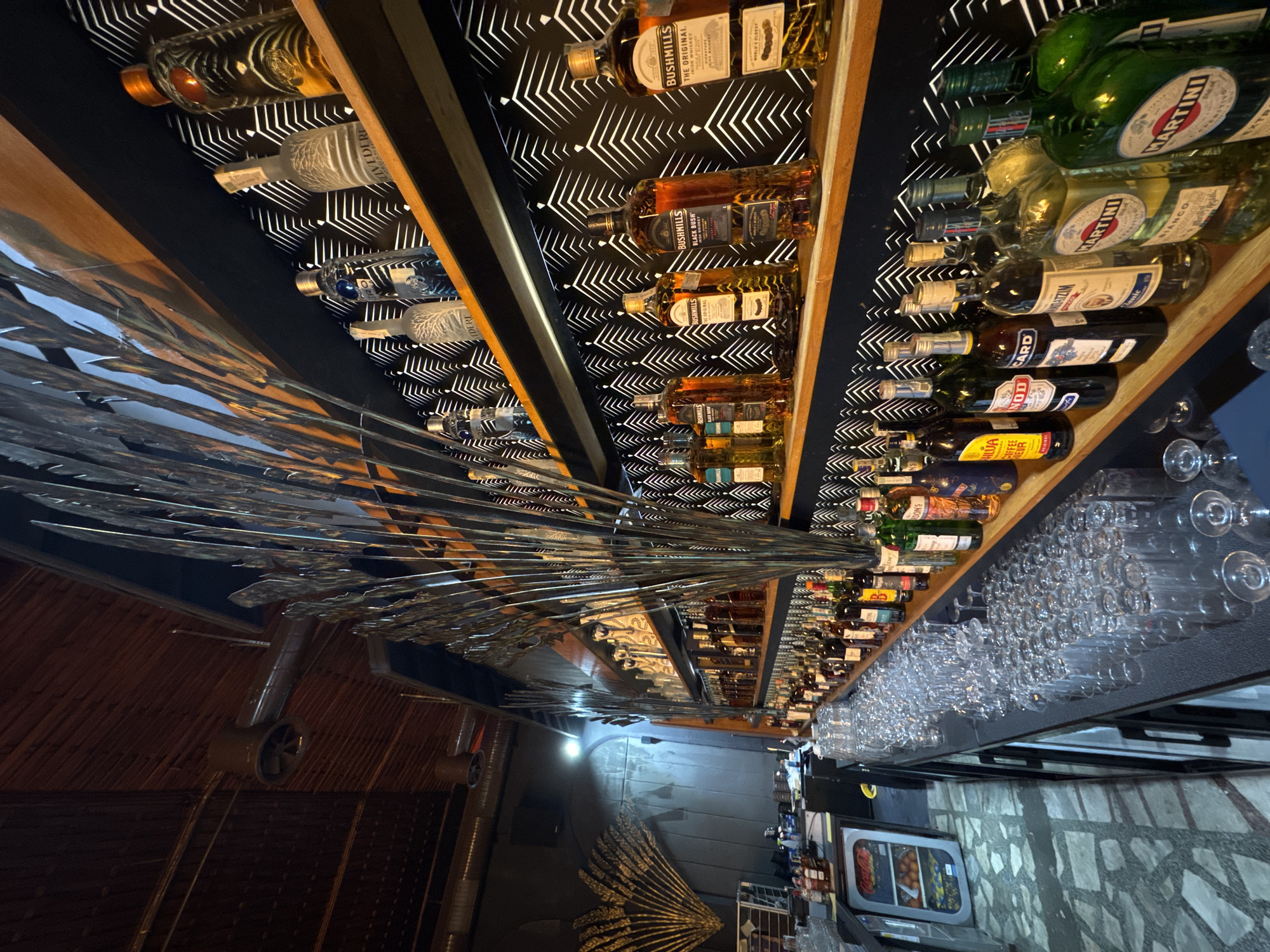 The image captures an indoor bar or liquor display area, likely located in Tsarevo, Bulgaria, viewed from a slightly upward angle and rotated 90 degrees clockwise. The main subjects are numerous bottles of various alcoholic beverages arranged on wooden shelves against a striking black background with a repeating white geometric pattern.
No people are visible, and no specific activity is taking place, but the setup implies a functional bar ready for service. On the left side of the frame, a large, intricate metallic sculpture or chandelier with many reflective, pointed elements dominates the space, reflecting light. Below it, a dark floor and ceiling fixtures are partially visible.
To the right, a long bar counter is meticulously lined with rows of clean, empty stemware and other glasses, indicating preparation for serving drinks. Further along the counter, a beverage cooler and a patterned floor can be seen. The lighting is artificial, illuminating the bottles and creating reflections on the metallic decor and glasses.
Visible text on the bottles, as confirmed by OCR, includes:
* Brands: BUSHMILLS ("THE ORIGINAL IRISH WHISKEY", "BLACK BUSH WHISKEY"), MARTINI ("BIANCO", "EXTRA"), BELVEDERE, Kahlúa (labeled as "COFFEE LIQUEUR"), Don's, and Nod Card.
* Other words: "WORLD'S OLDEST".
The scene suggests an evening or nighttime setting for a bar, showcasing its extensive selection of spirits and readiness for patrons.