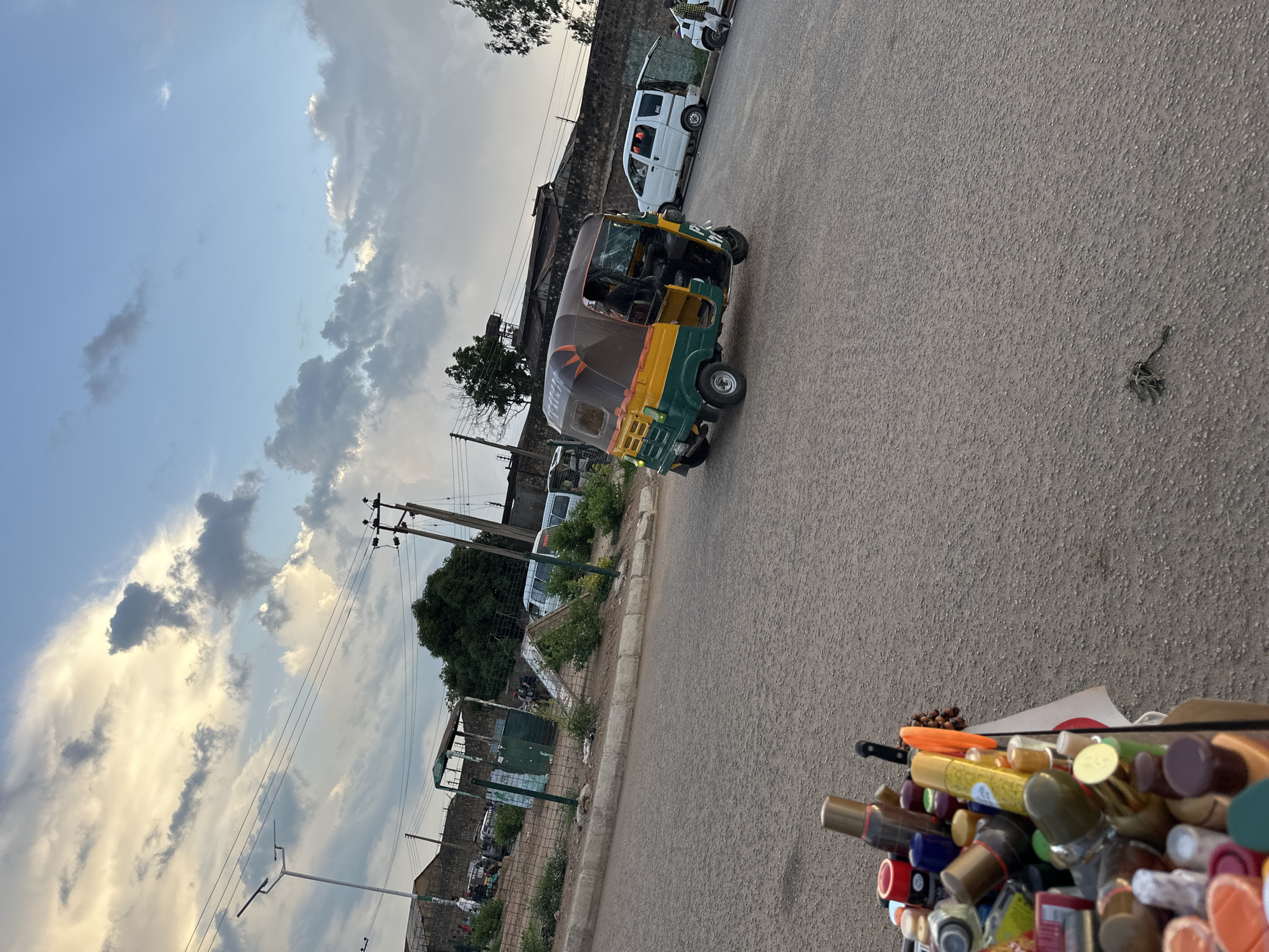 In Bukuru, Nigeria, a street scene unfolds with a yellow and green auto-rickshaw in motion on an asphalt road. The auto-rickshaw is occupied by a person in the driver's seat. To the right of the auto-rickshaw, a white van with its rear doors open is parked, and another white van is visible further down the road. A man in a white shirt and black pants stands near the parked white van. On the left side of the road, a cart or display is filled with various bottles, including perfume bottles and other containers. In the background, buildings with brick walls and green fencing are present. A street light stands on the left side of the road, and utility poles with wires are visible throughout the scene. The sky above is partly cloudy with dramatic cloud formations.