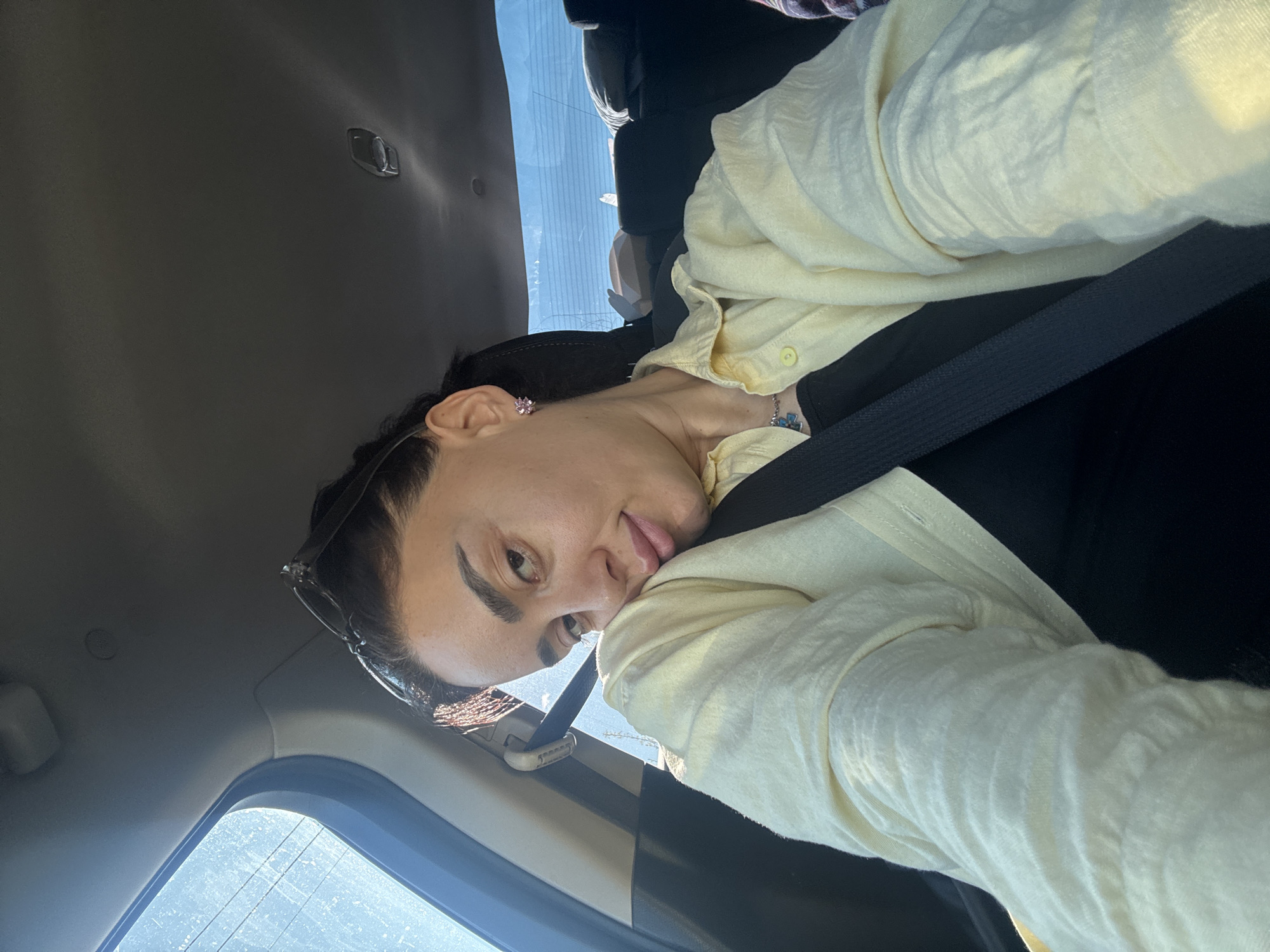 A person is in a car. The person is wearing a black top and a light yellow button-down shirt. They have a seatbelt on. Sunglasses are perched on top of their head. The car is likely in Tbilisi, Georgia, based on the location information. The car is in transit during daylight hours.