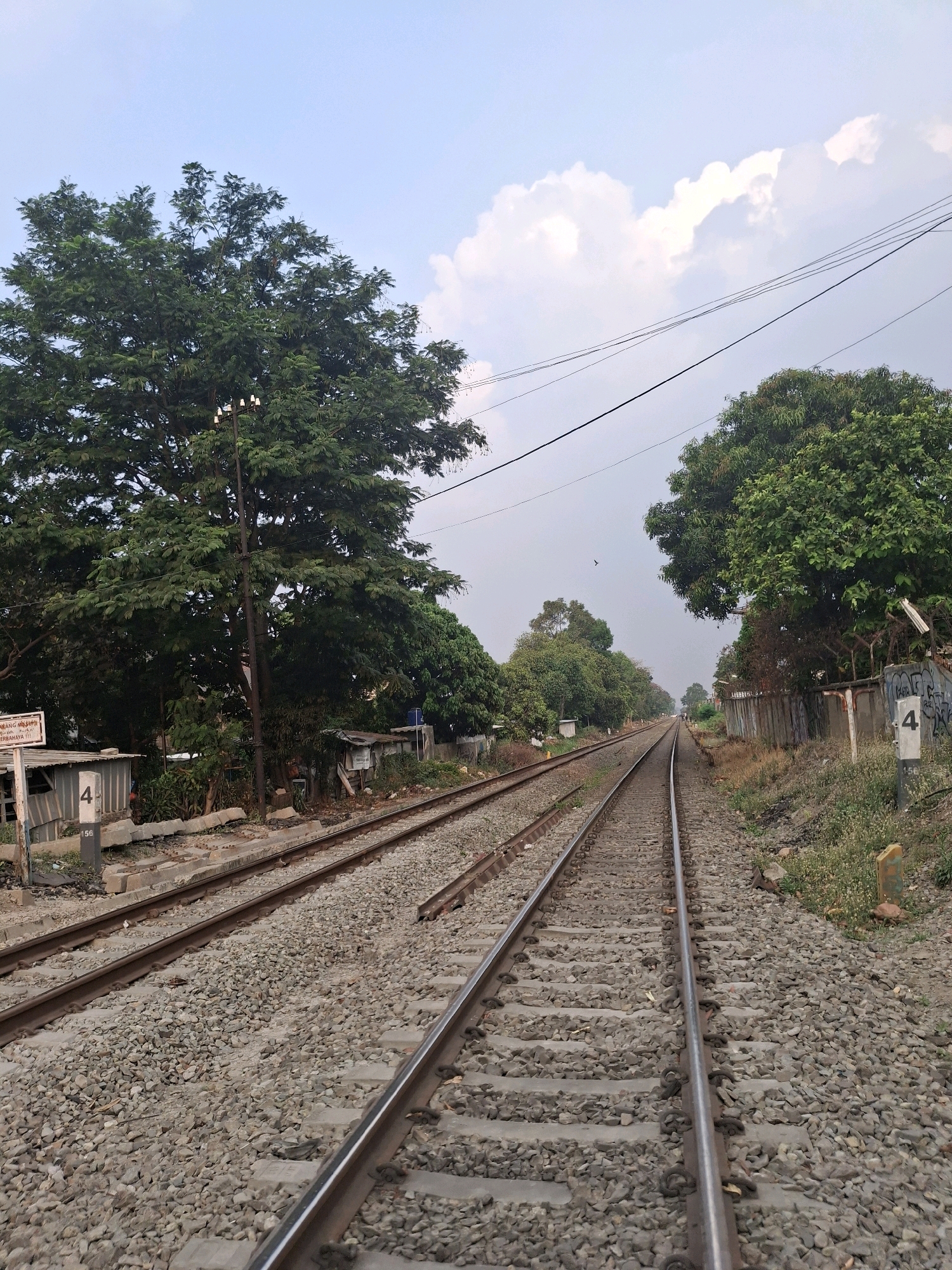 A set of parallel train tracks runs through a rural or semi-urban landscape under a partly cloudy sky. The tracks are made of gravel and are flanked by grassy areas and vegetation. On the left side of the tracks, there are some low-lying structures, a large tree, and a utility pole with lights. On the right side, there is a fence with graffiti and a concrete marker with the number "4". The image appears to be taken from ground level, looking down the length of the tracks as they recede into the distance. There are no people or vehicles visible in the frame. The overall impression is one of an industrial or transportation infrastructure in a natural setting.