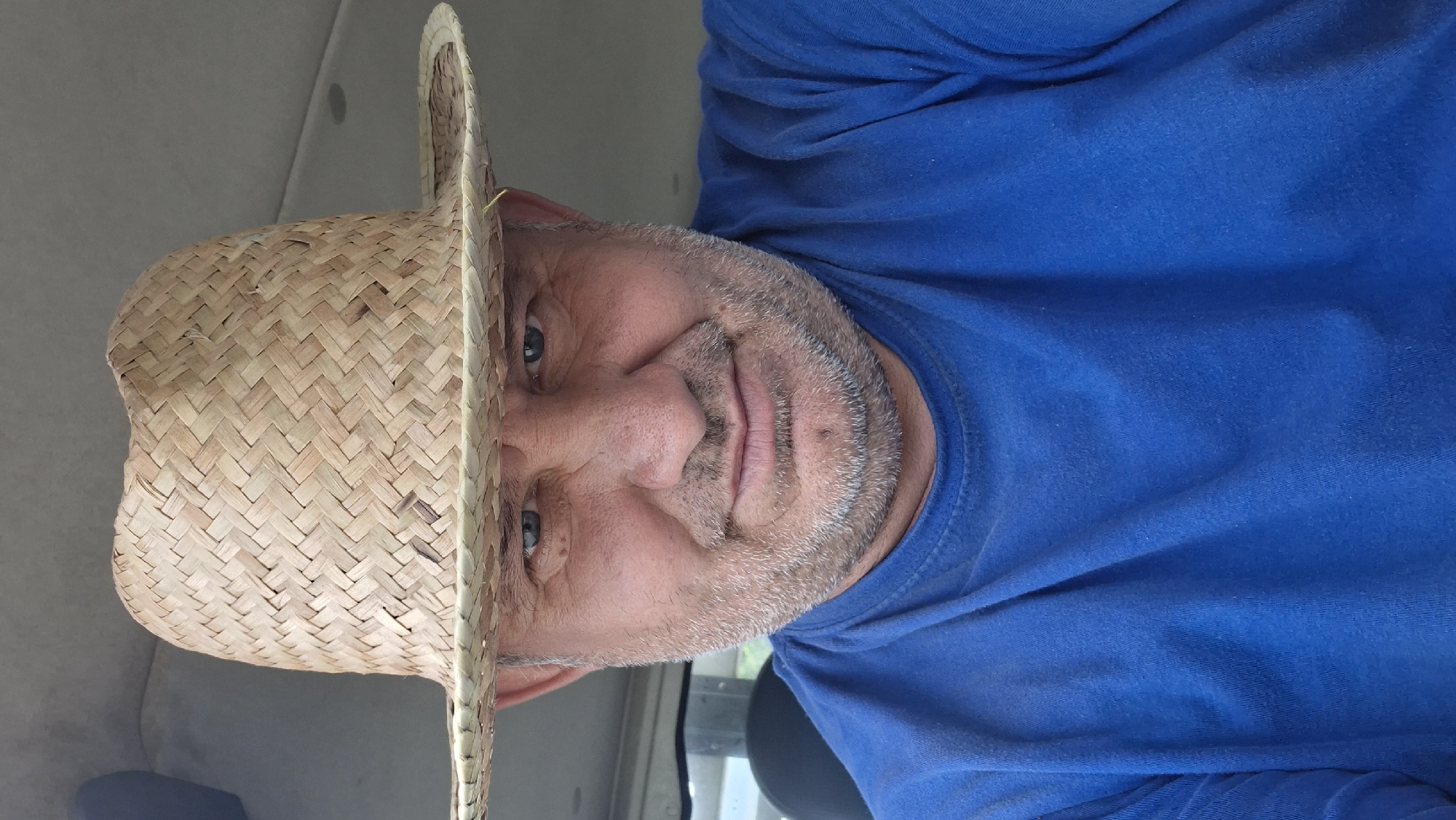 A man wearing a straw hat and a blue t-shirt is captured in a close-up selfie. The man has light blue eyes and a beard. The background suggests an indoor setting, possibly a vehicle, with a grey ceiling and a hint of dark upholstery on the left. The lighting is natural and comes from the front, casting soft shadows on the man's face. The image's context does not provide specific information about its location in Hîncești, Moldova.