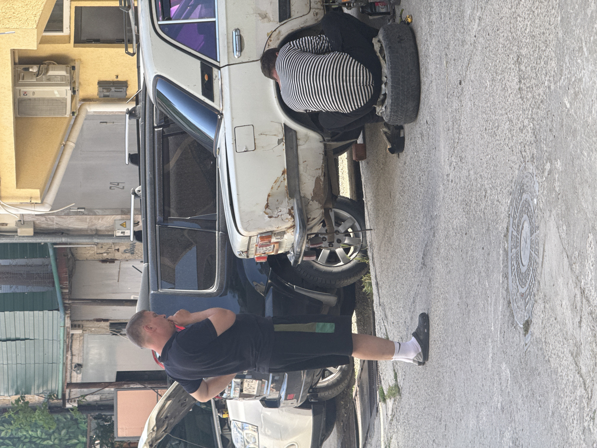 A medium, vertical shot depicts two men working on the roadside on a row of cars in the city of Unknown, Unknown, under daylight conditions. One man is dressed in a black-and-white striped shirt and dark pants. He is bent at the waist near the front tire of a white vehicle, possibly engaged in repairs or maintenance. Adjacent to him, resting on the ground, is a black tire. Standing next to the vehicle, on the sidewalk, is a man dressed in a black short-sleeved shirt and black shorts. He is standing and appears to be occupied with something. Behind these two men are several other parked vehicles in a row. The backdrop reveals a building with a yellow exterior, windows, and an air conditioning unit, followed by a tall grey wall with the number "24" painted on it. The sidewalk is a light gray color, and the overall environment suggests an urban setting.