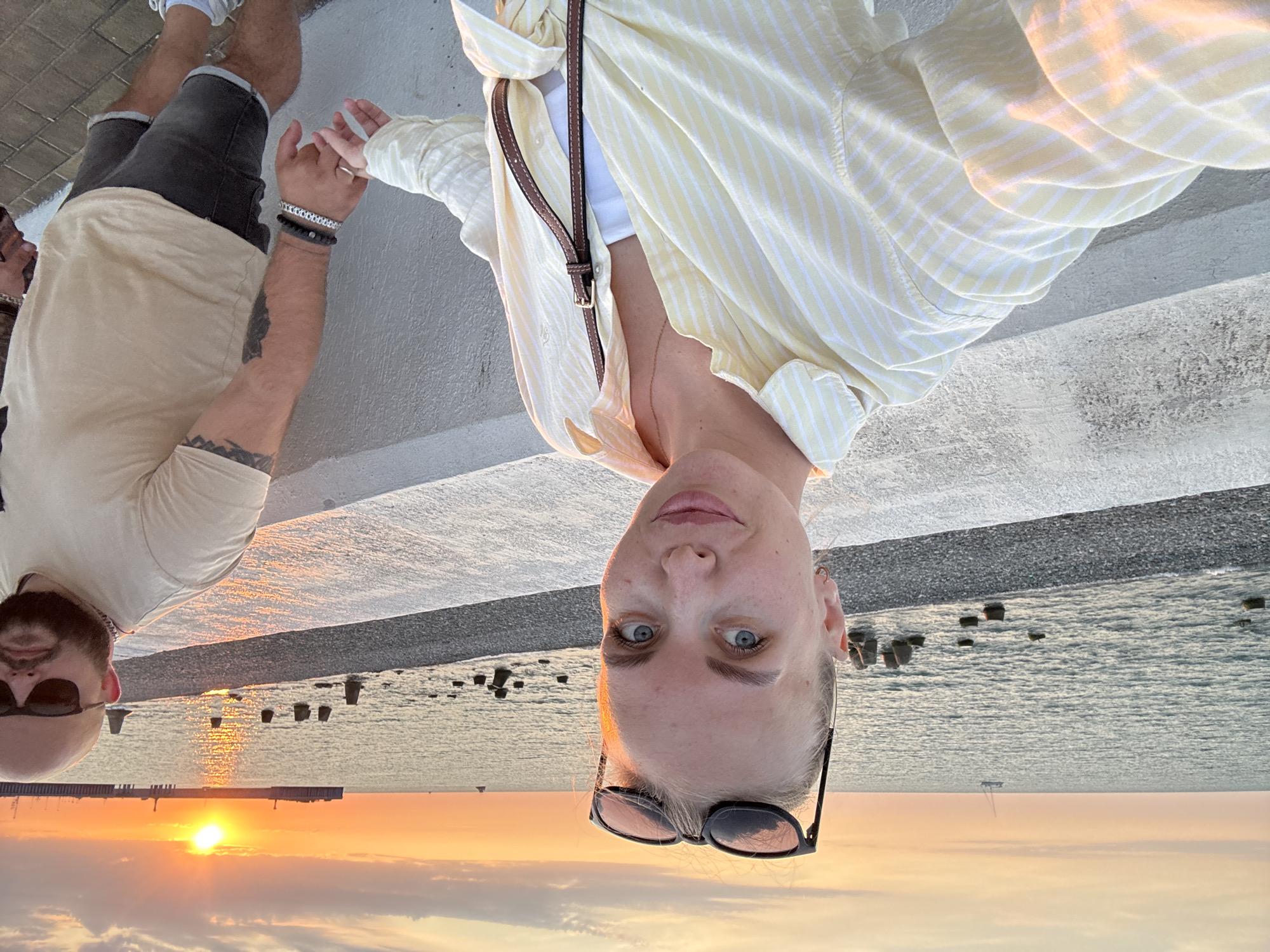 The image captures a couple holding hands on a promenade at sunset in Sirius, Russia. The woman, in the foreground, is wearing a light yellow striped shirt over a white top and sunglasses pushed up onto her head. She has blonde hair and blue eyes. The man, to her right, is wearing a beige t-shirt and dark denim shorts, with sunglasses and jewelry on his wrist. They are walking along a concrete barrier that overlooks a pebble beach and the Black Sea. The sun is setting on the horizon, casting a warm glow over the scene, with a sailboat visible in the distance and a pier with lights.