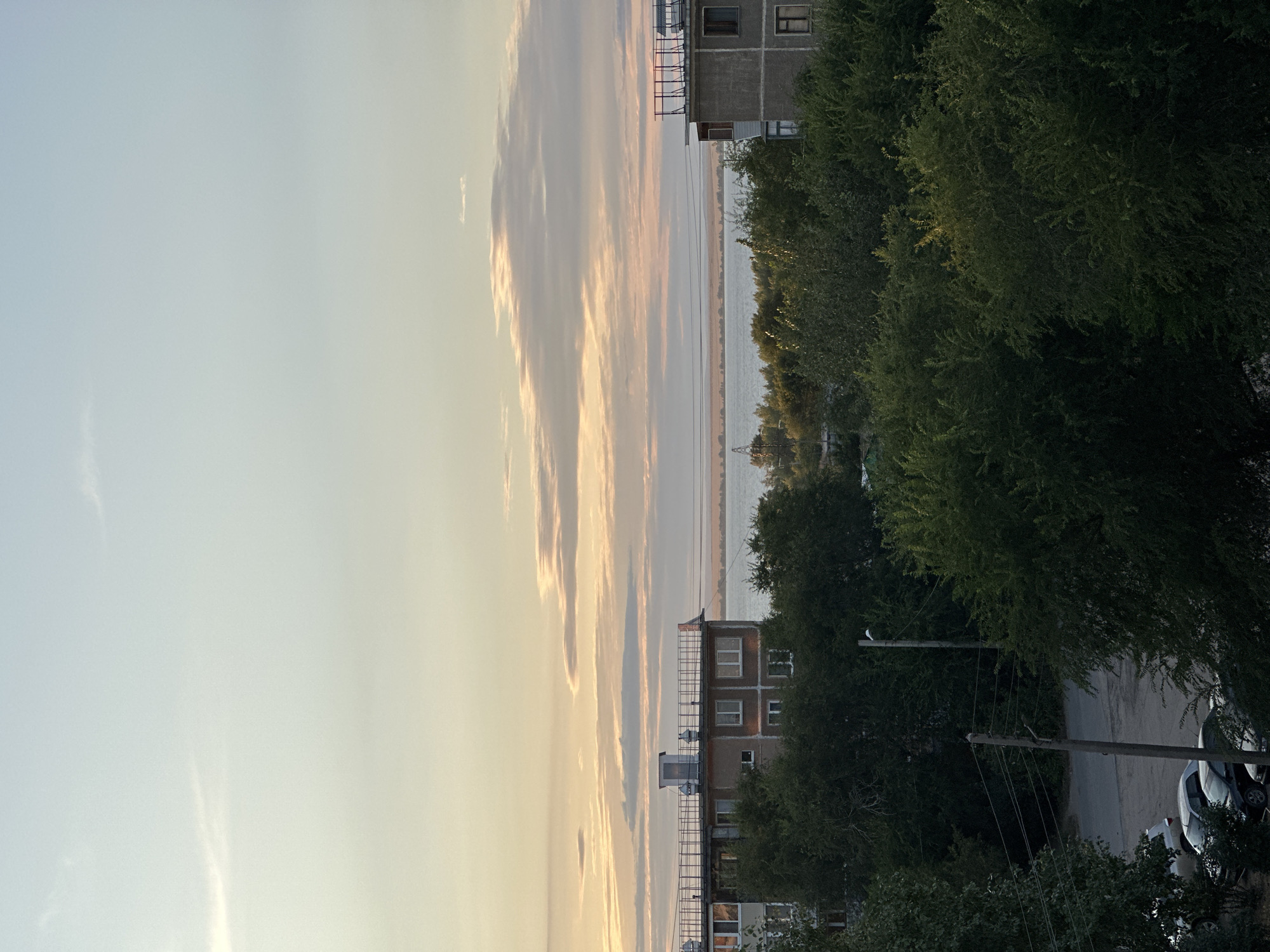 The image captures a tranquil scene in the city of Energetik, Russia, likely during the late afternoon or early evening, given the soft, warm light illuminating the sky. The sky is a canvas of muted blues and yellows, with wispy clouds stretched horizontally across the horizon, hinting at the setting sun.
Dominating the middle ground are two apartment buildings, characteristic of Soviet-era architecture. The building on the left appears to be a multi-story structure with a flat roof equipped with railings and a small utility structure. Its facade is a neutral beige color, punctuated by rows of windows. The building on the right, also multi-story, has a grey, concrete facade and similar window placements. Both buildings are partially obscured by the lush, dense foliage of trees that front them.
Beyond the buildings and trees lies a wide body of water, likely a river or a lake, stretching towards a distant, hazy shoreline. The water reflects the soft colors of the sky, adding to the serene atmosphere. The landscape beyond the water is indistinct due to the distance and atmospheric conditions.
In the foreground, the dense greenery of trees dominates, creating a natural screen that frames the view of the buildings and the water. The trees are full and vibrant, suggesting a season of growth, possibly summer or early autumn. Peeking through the trees on the left side of the image, a glimpse of a paved area can be seen with several parked cars. A wooden utility pole with wires stretches vertically through this area, a common sight in urban environments. No people are visible in the frame, contributing to the sense of stillness and quiet. The overall impression is one of a peaceful urban landscape integrated with nature.