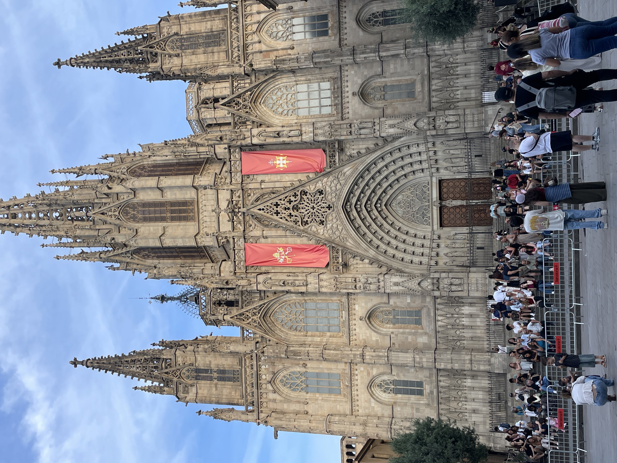 This image captures the grand Gothic facade of the Cathedral of Barcelona (Catedral de la Santa Cruz y Santa Eulalia de Barcelona) in Barcelona, Spain, under a partly cloudy blue sky. The cathedral, constructed from light-colored stone, features three prominent spires, numerous buttresses, ornate tracery, and multiple arched windows. The central section is dominated by a large, intricately carved arched portal leading to double wooden doors. Above the main entrance, a large rose window is visible, surrounded by detailed sculptural work. Two large red banners hang from the upper-central facade; the left banner displays a golden cross within a circular emblem, while the right banner features the papal tiara and crossed keys.
At the base of the cathedral, a wide set of stone steps ascends to the main entrance. A significant crowd of over one hundred individuals is present in the foreground and on these steps. Many people are seated on the steps, observing their surroundings or looking at their phones. Others stand in the paved plaza area directly in front of the cathedral, positioned both behind and in front of temporary metal barriers, some bearing the text "Barcelona" and "Ajuntament de Barcelona."
The individuals are a diverse group of adults, teenagers, and some children, dressed in casual summer attire such as t-shirts, shorts, and jeans. In the immediate foreground, a woman in a white top and blue jeans crouches on the paved ground, facing towards the right. Nearby, a young boy in a blue t-shirt and shorts stands looking towards the right. Further back, a man with a grey backpack stands facing the cathedral, while a group of people to his right also face the cathedral, some appearing to be taking photos. The scene suggests a public gathering or tourist activity in front of the landmark. A large green tree is visible on the left side of the frame. The overall lighting is bright and natural, indicating daytime conditions.