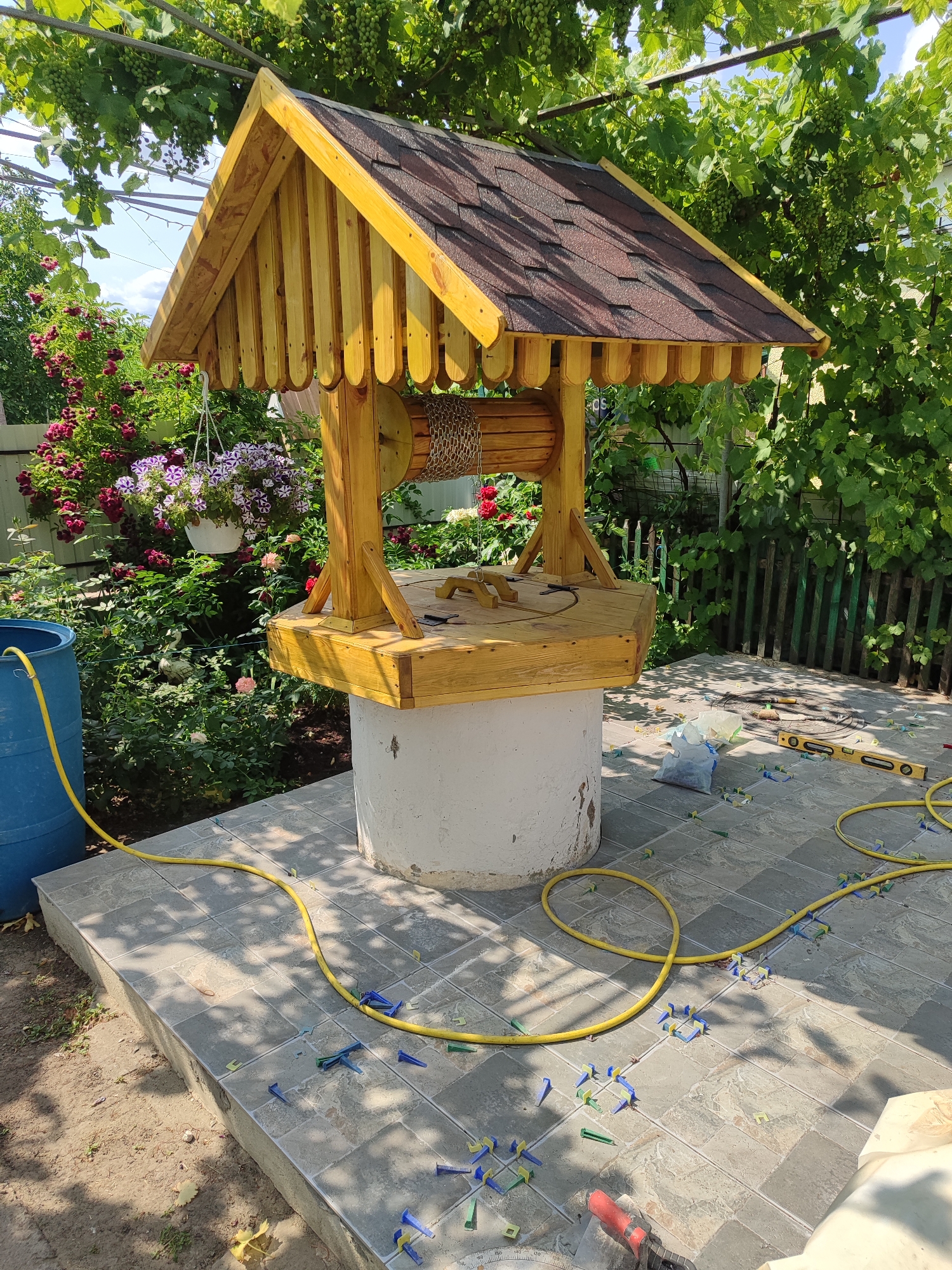 In Novosamarka, Ukraine, a wooden well with a shingled roof stands on a white circular base in a garden. A yellow hose is connected to a large blue barrel to the left and winds across the tiled ground, which is scattered with blue and green tile spacers. The well's wooden platform holds various tools, including a level and a small red power tool. Behind the well, a fence and lush green foliage with blooming red roses and potted flowers create a natural backdrop. Sunlight filters through the leaves, casting shadows on the tiled area.